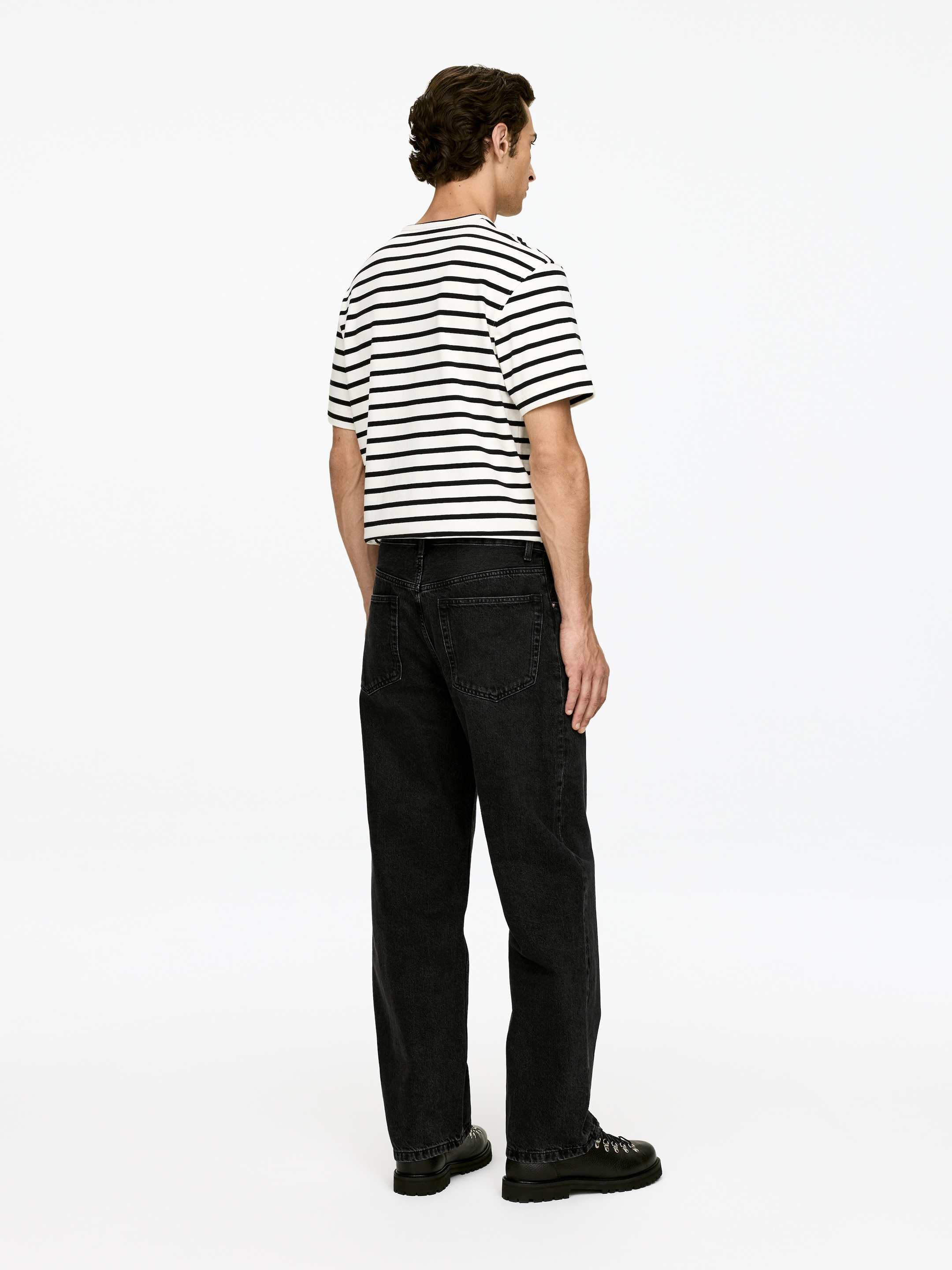 Grösseres Bild ansehen: Ein weißes kurzärmeliges T-Shirt mit horizontalen schwarzen Streifen wird getragen und weist einen leicht verkürzten Saum auf. Es wird mit dunklen, weiten Jeans und schwarzen Stiefeln mit dicken Sohlen kombiniert, alles aus einer Rück- und leicht seitlichen Ansicht präsentiert.