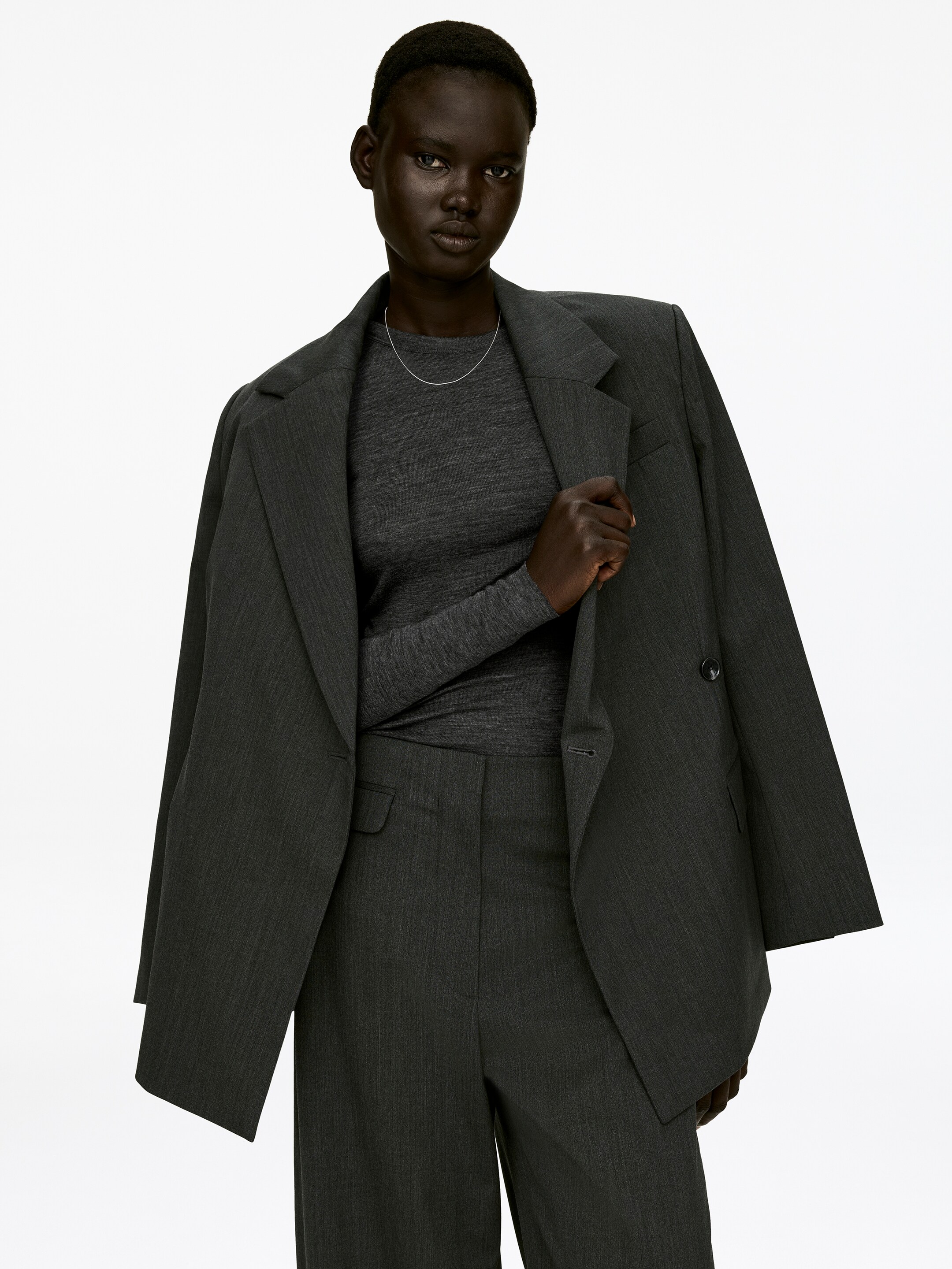 Größeres Bild anzeigen: Ein dunkelgraues, eng anliegendes Merinowoll-Top mit Rundhalsausschnitt und langen Ärmeln wird von dem Model getragen. Das Top erscheint leicht transparent und wird unter einem offenen grauen Blazer geschichtet, während eine dünne silberne Halskette auf seinem Ausschnitt ruht.