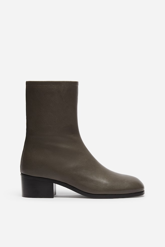 Ein einzelner dunkelolivgrauer Leder-Stiefelette ist in Seitenansicht dargestellt und verfügt über einen enganliegenden Schaft sowie eine abgerundete Spitze. Der Stiefel ist mit einem niedrigen schwarzen Blockabsatz und einer dünnen schwarzen Sohle gestaltet, was die minimalistische Silhouette betont. - 1