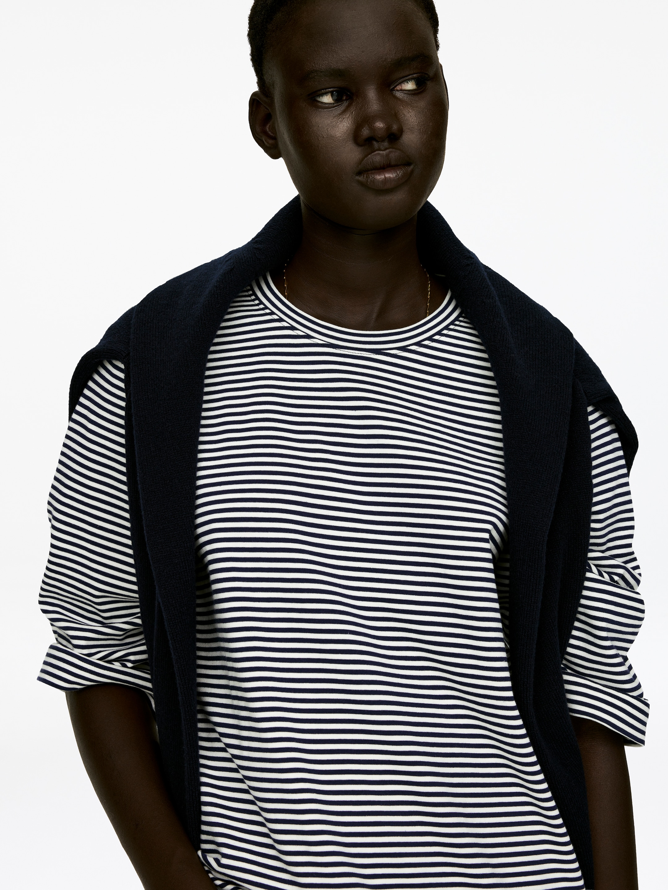 Agrandir l'image: Un T-shirt ras du cou, confectionné en jersey de coton épais et frais avec des rayures horizontales bleu marine et blanches et des manches de longueur 3/4, est porté par le mannequin. Les manches sont actuellement retroussées, et un pull foncé est drapé sur les épaules du mannequin, reposant sur le T-shirt.