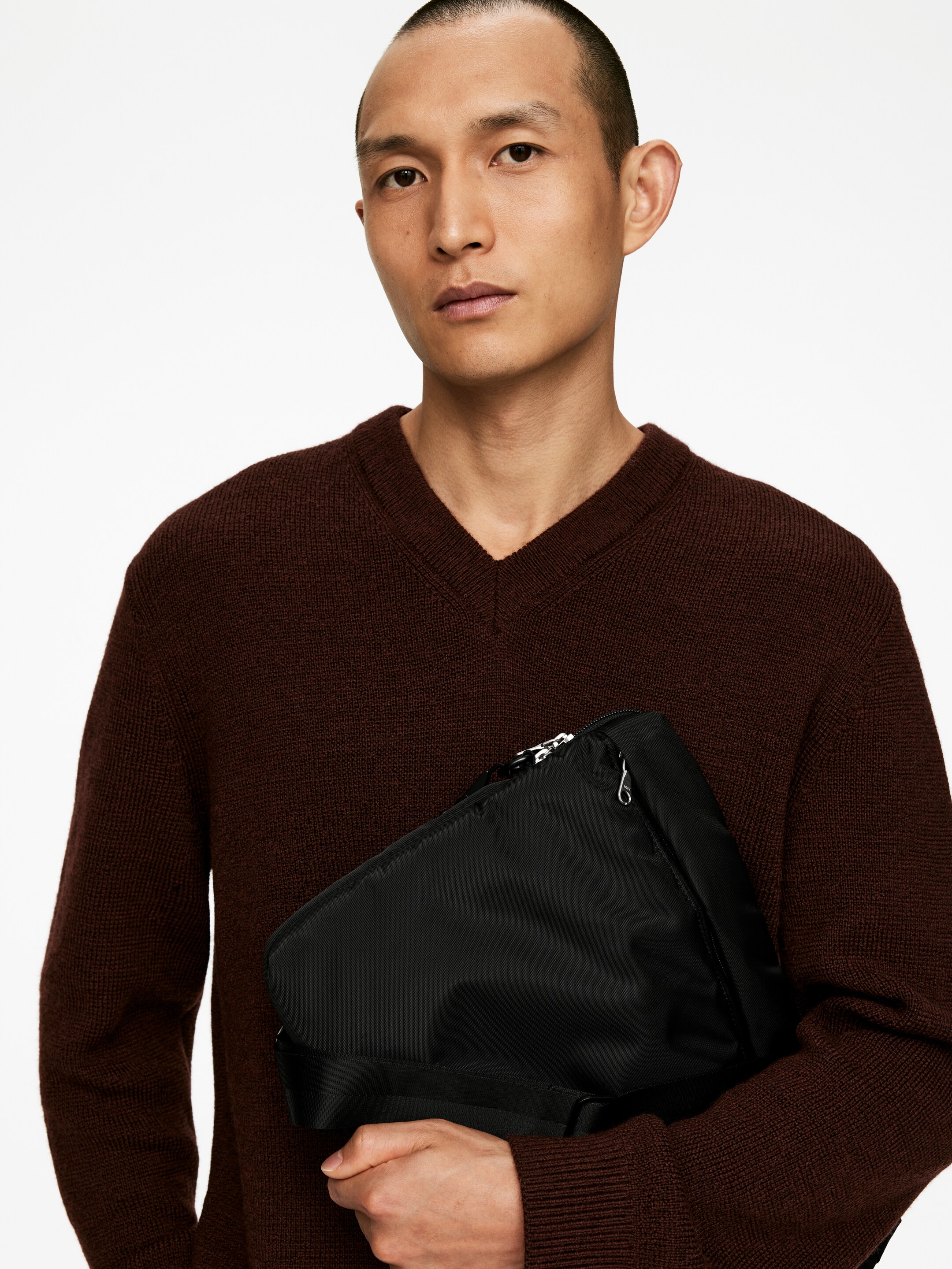 Agrandir l'image: Un sac noir est tenu par une personne, son corps principal reposant contre leur torse et légèrement incliné loin du spectateur. Son tissu noir doux et froissé est visible, avec une fermeture éclair de couleur argentée le long de son bord supérieur et une large sangle noire le long de sa partie inférieure avant.