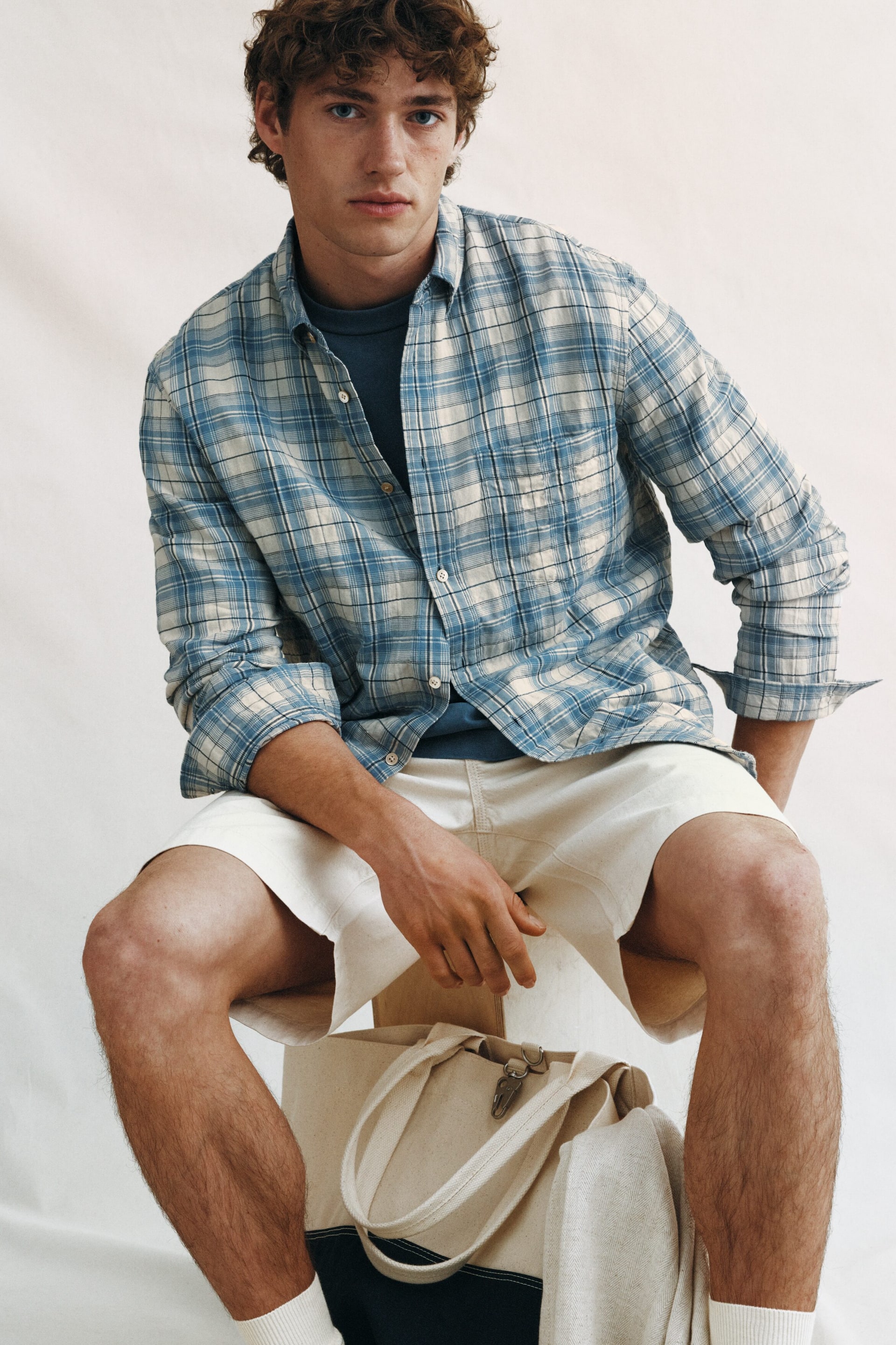 View larger image: A blue and cream checked button-down shirt, featuring rolled-up long sleeves and a single chest pocket, is being worn open over a dark blue crew neck t-shirt. Cream-coloured shorts with visible stitching are also being worn.