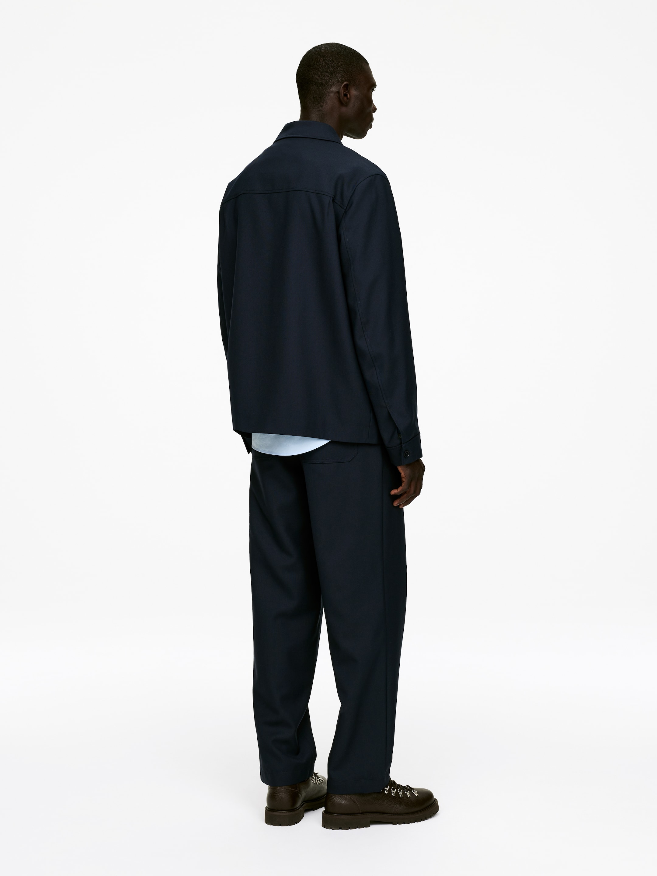 Agrandir l'image: Une veste bleu foncé avec un col pointu et un empiècement horizontal dans le dos, ainsi qu'un pantalon assorti à coupe décontractée, sont présentés de dos, légèrement en angle. Un sous-vêtement bleu clair est porté sous la veste. Des bottes marron foncé à semelles crantées sont stylisées avec le pantalon.