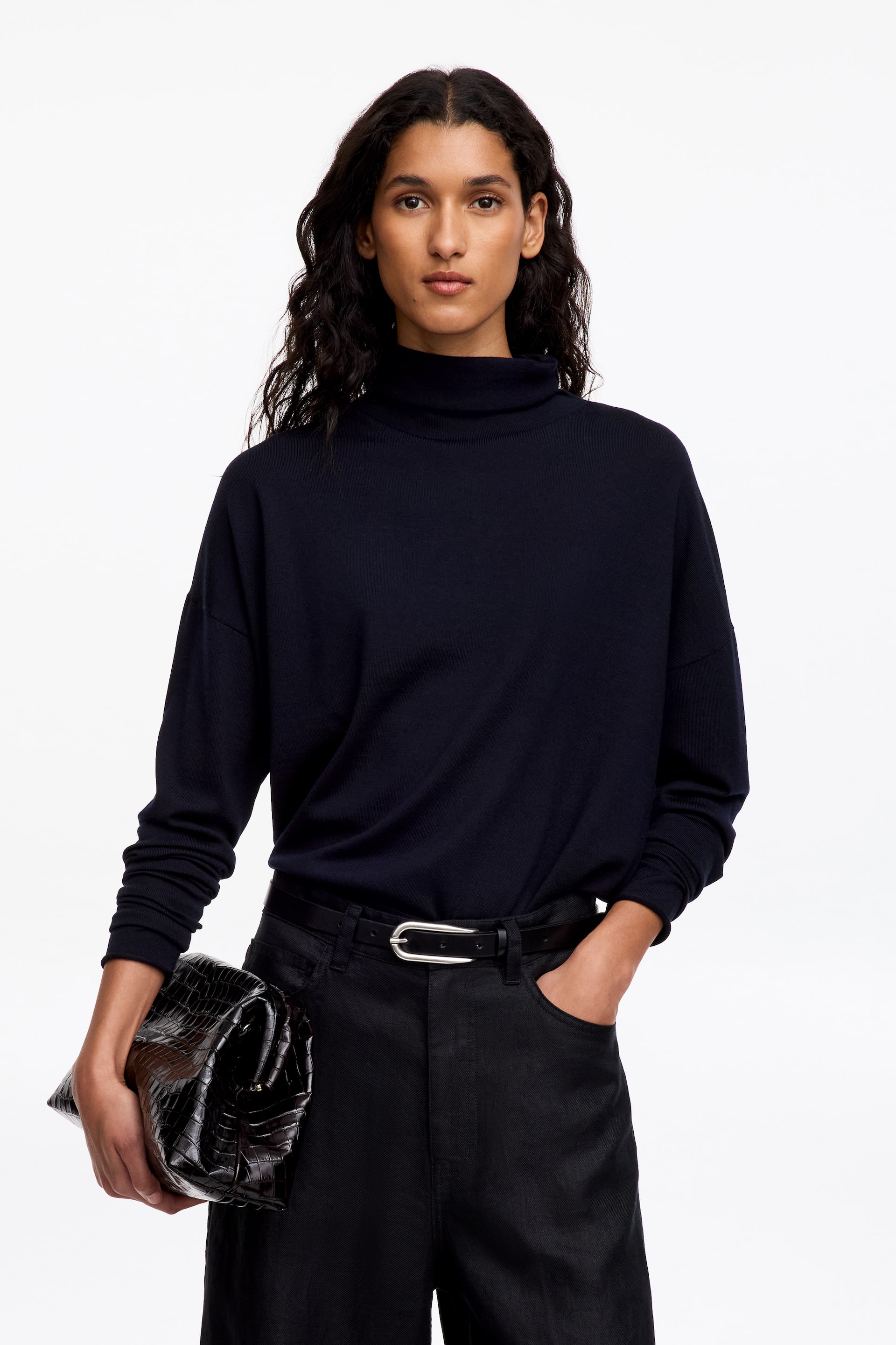 Agrandir l'image: Un pull en maille bleu marine foncé, doté d’un col roulé souplement drapé et de manches longues décontractées, est porté par le mannequin. Le pull est rentré dans un pantalon noir maintenu par une ceinture à boucle argentée, tandis que le mannequin tient une pochette noire texturée.