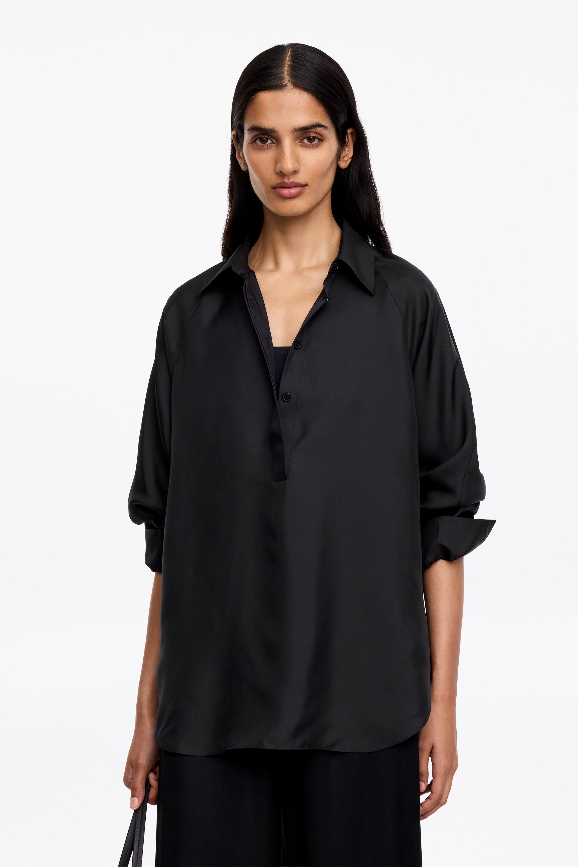 Visualizza l'immagine ingrandita: Una camicia nera con colletto presenta un mezzo abbottonatura con tre bottoni e maniche raglan, arrotolate fino agli avambracci. È tagliata con una vestibilità rilassata, mostrando una maglietta scura sotto il colletto aperto.