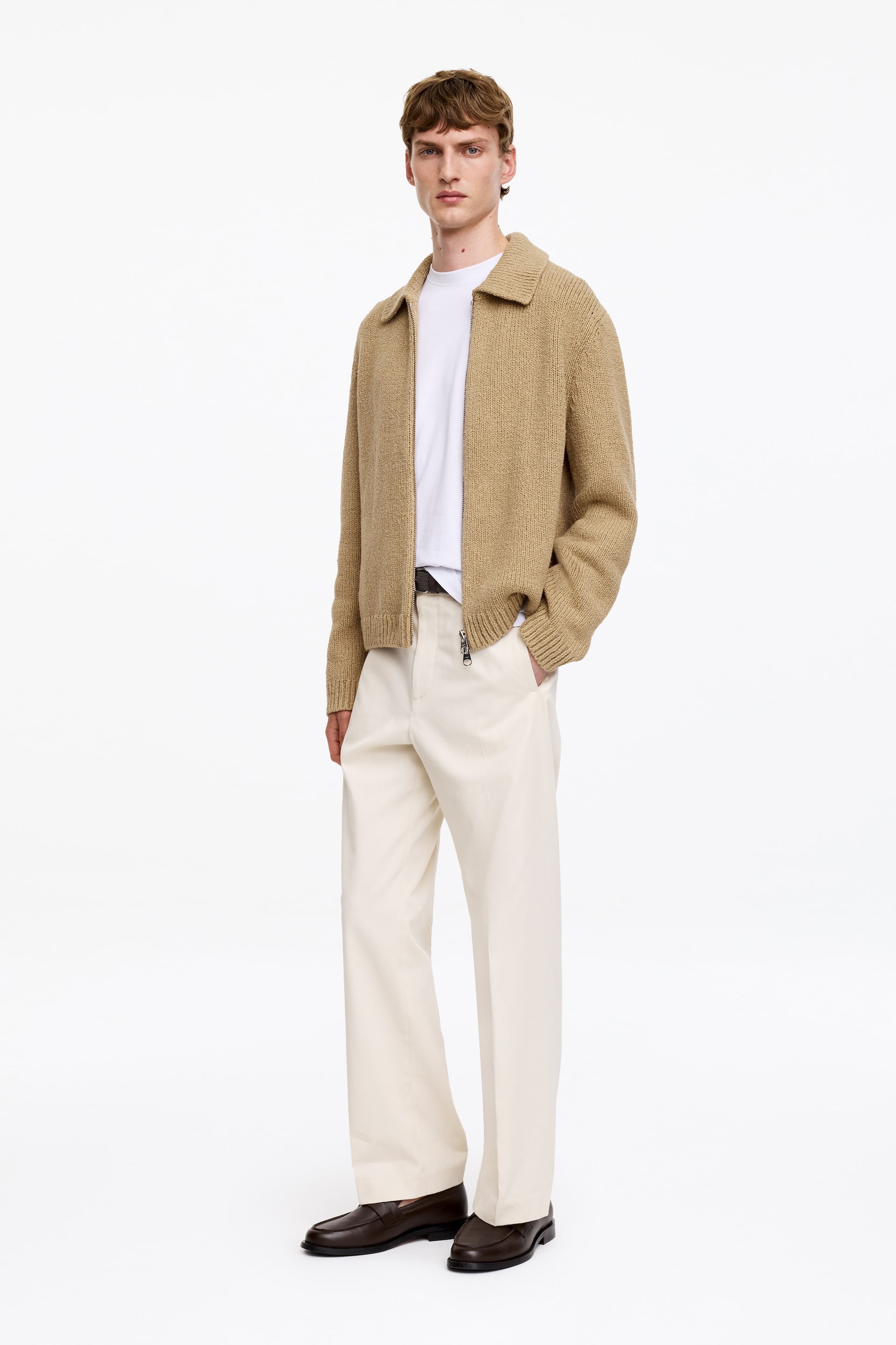 Agrandir l'image: Une veste zippée en tricot texturé beige est portée à un léger angle frontal, mettant en valeur son col standard, sa fermeture éclair intégrale, et ses poignets et ourlet côtelés. Elle est superposée à un t-shirt blanc à col rond et associée à un pantalon ample crème, une ceinture foncée et des mocassins marron foncé.
