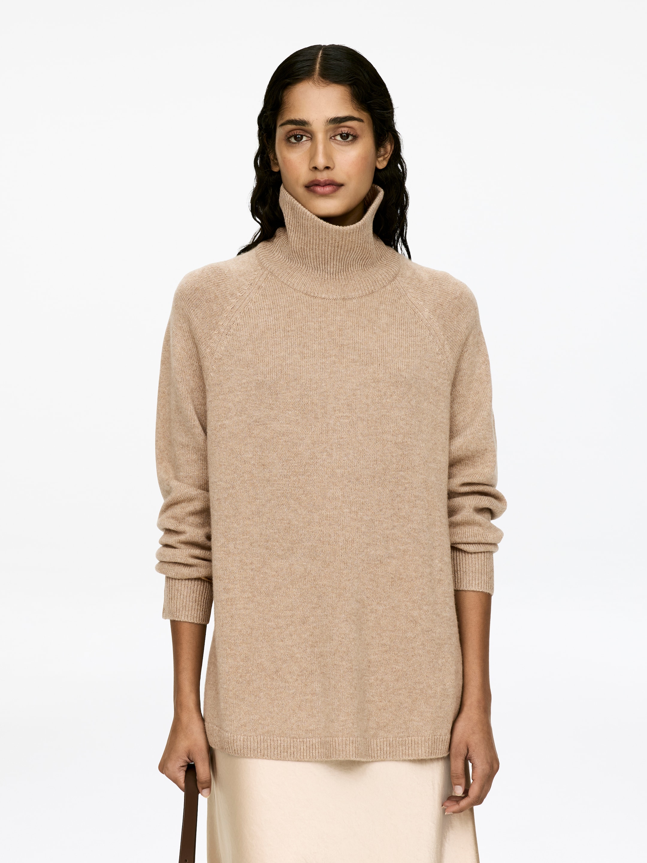 Visa större bild: En beige polotröja, tillverkad av kashmir och merinoull, bärs av modellen. Tröjan har raglanärmar och ribbstickade detaljer på hals, muddar och fåll, och är lager på lager över en ljusfärgad kjol.