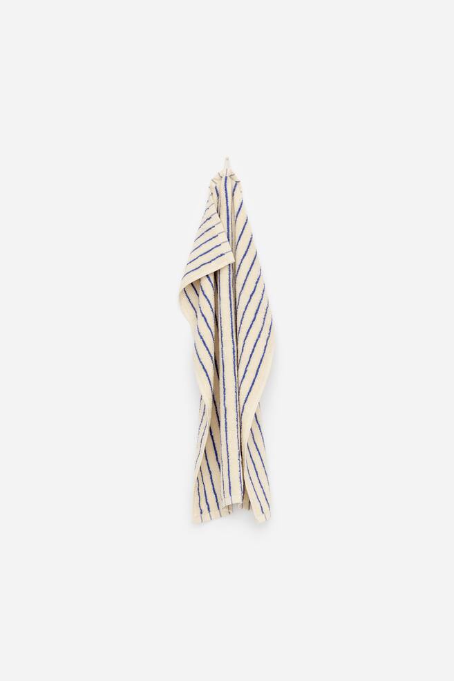 A hand towel crafted from soft cotton terry is neatly folded and hanging, showcasing a cream background adorned with vertical blue stripes. A hanging loop is evident at the top. - 1