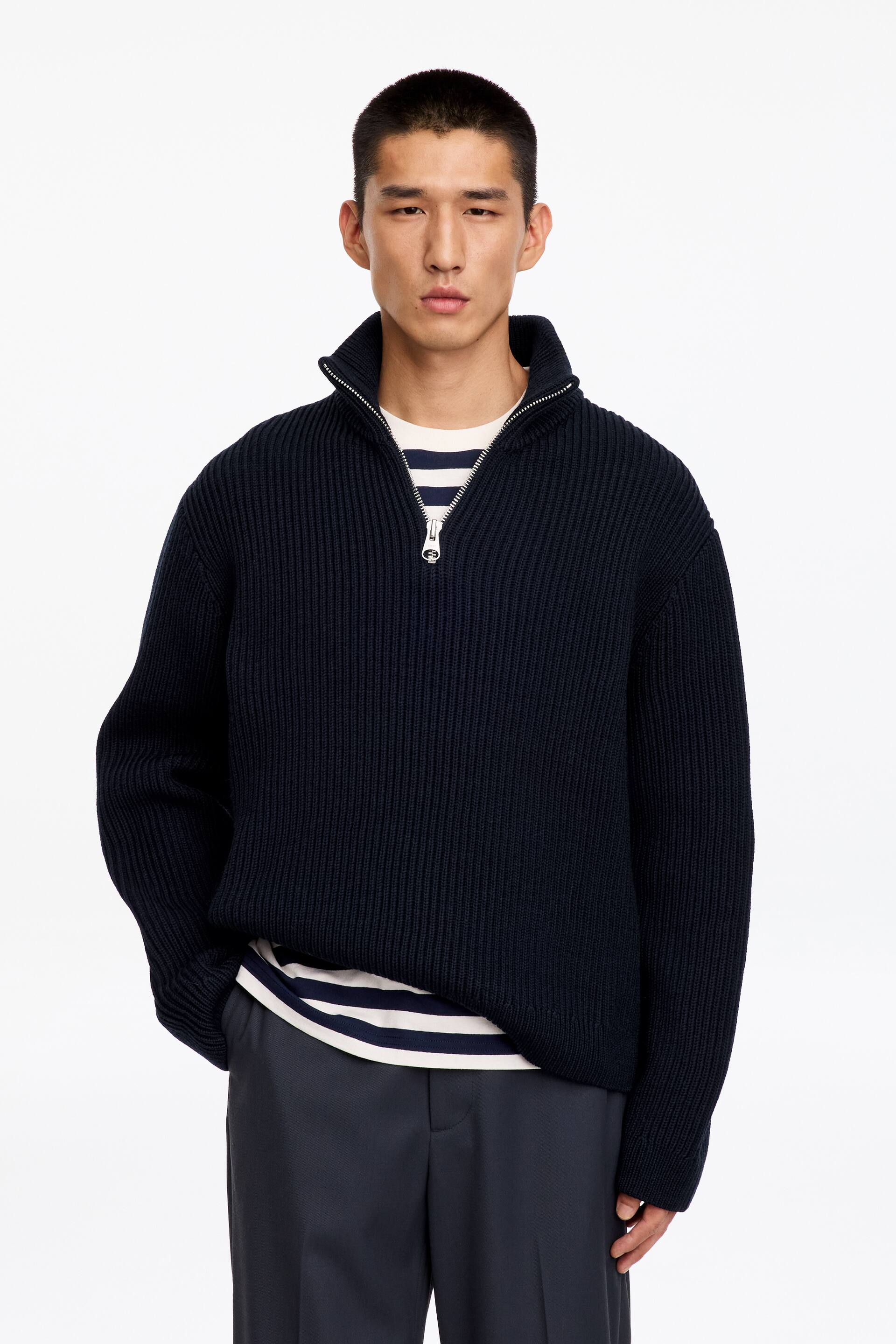 View larger image: A navy blue half-zip knitted jumper in a full cardigan stitch is being worn by the model, featuring a ribbed funnel collar, cuffs and hem. The jumper is layered over a navy-and-white striped T-shirt and paired with dark trousers.