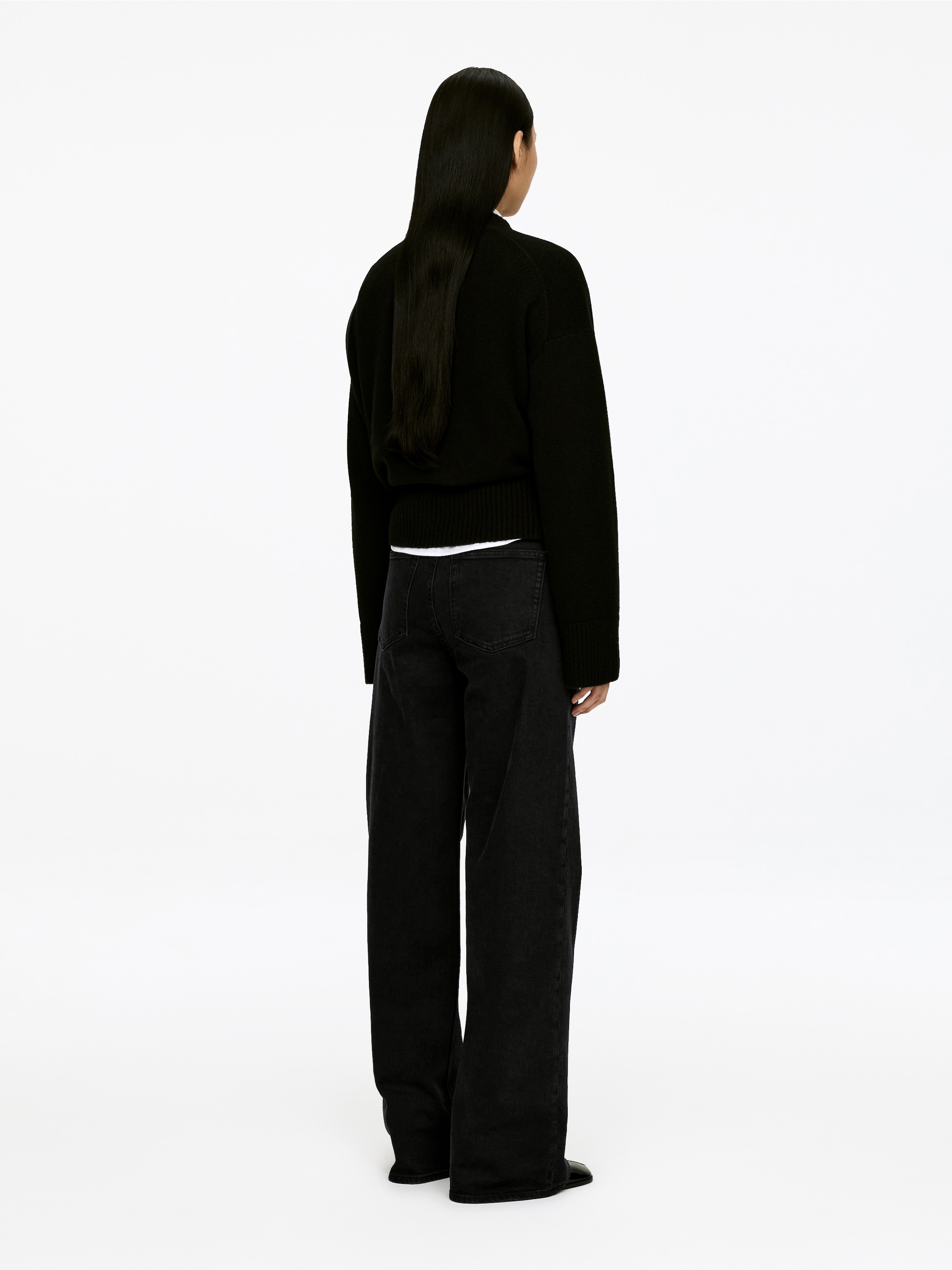 Größeres Bild ansehen: Ein schwarzer Pullover und schwarze Jeans mit weitem Bein werden aus der Rückansicht präsentiert. Der schwarze Pullover mit überschnittenen Schultern sowie gerippten Bündchen und Saum wird über einem weißen T-Shirt getragen. Die schwarzen Jeans aus Denim haben eine entspannte, weite Passform.