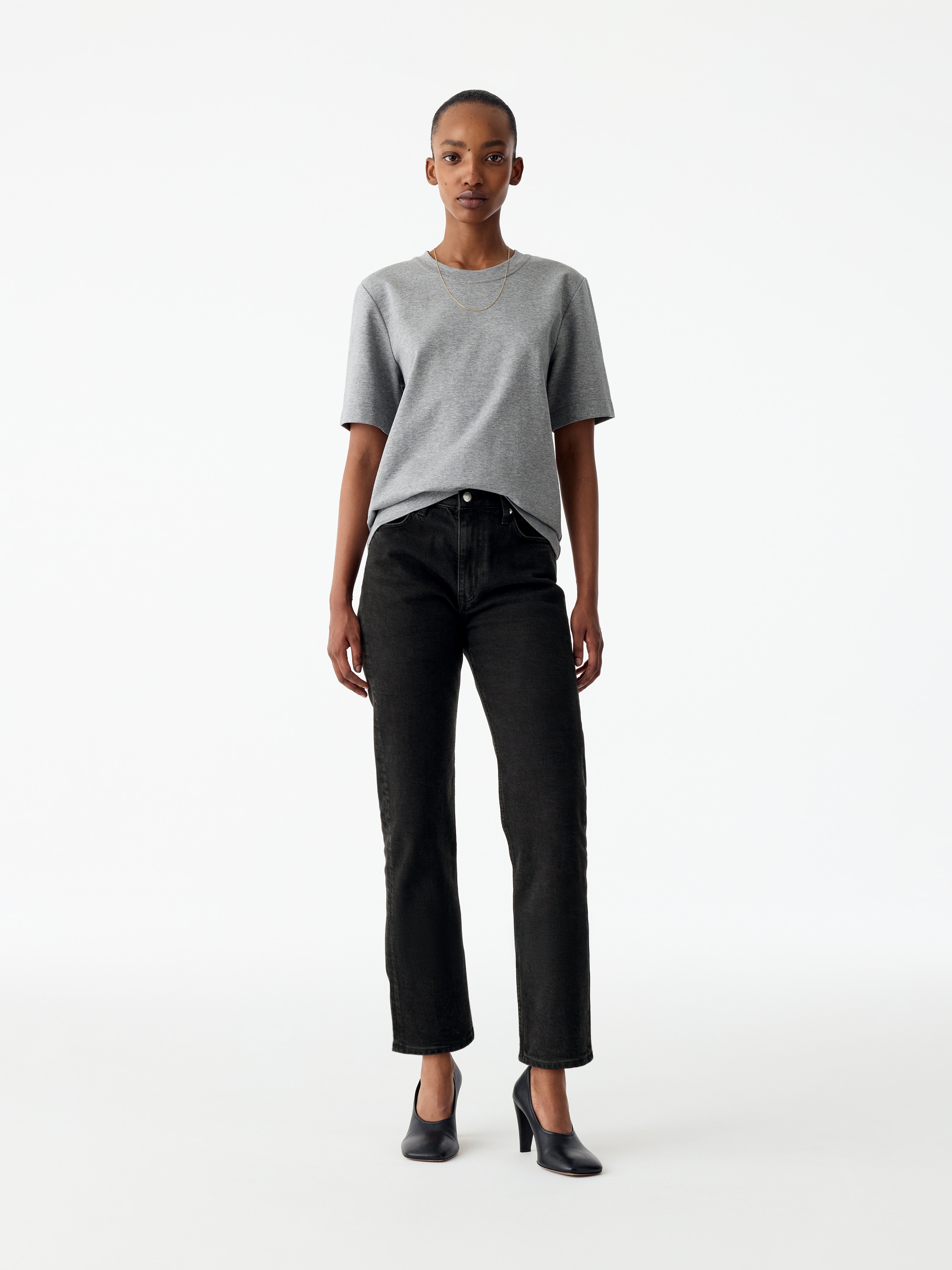 Ein Paar schwarze Jeans aus Denim wird flach präsentiert, mit der Vorderseite nach vorne. Sie verfügen über eine mittelhohe Taille, einen sichtbaren Reißverschluss und zwei Fronttaschen mit metallischen Nieten. Die Beine erscheinen schlank, schmal zulaufend und gekürzt.