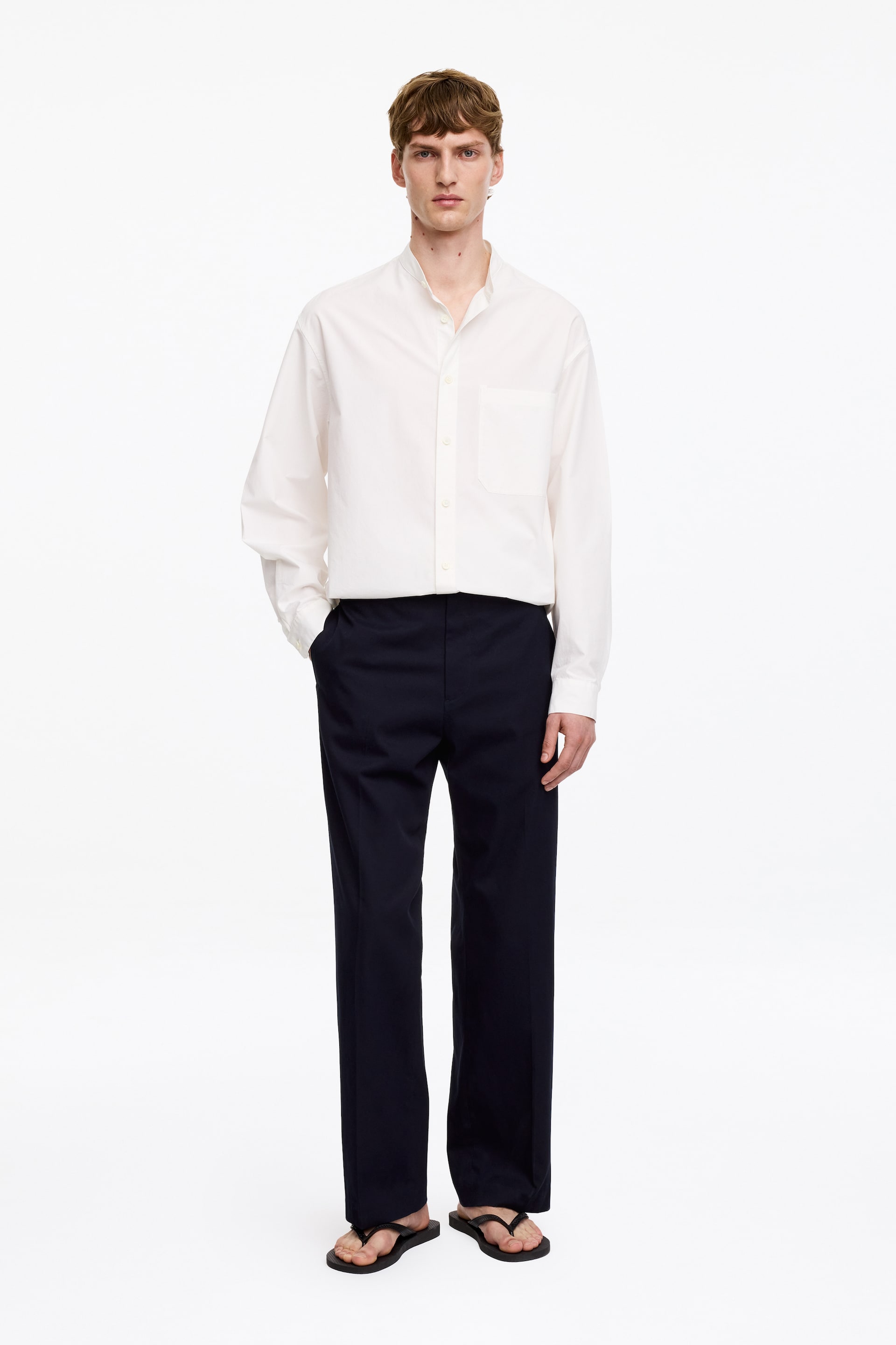 Grotere afbeelding bekijken: Donkerblauwe katoenen twill broek, met een relaxte, rechte pasvorm en een zijnaadzak, wordt door het model gedragen met een ingestopt wit overhemd. De linkerhand van het model is in de zak gestoken.