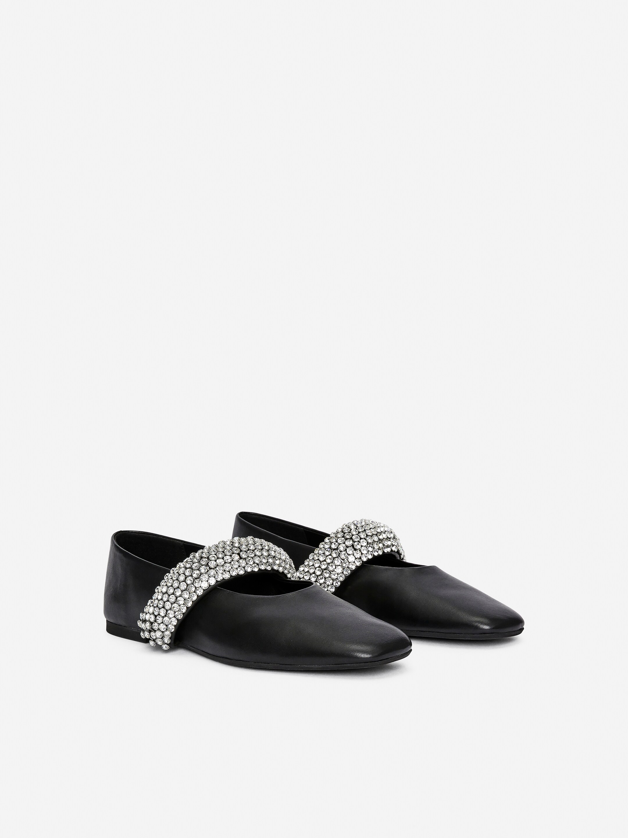 Größeres Bild ansehen: Ein schwarzer flacher Schuh ist auffällig auf der linken Seite zu sehen, seine leicht spitze Zehe zeigt nach rechts und offenbart einen breiten Ristgurt. Der Gurt ist stark mit mehreren Reihen funkelnder klarer Strasssteine verziert, während ein zweiter ähnlicher Schuh teilweise dahinter sichtbar ist.