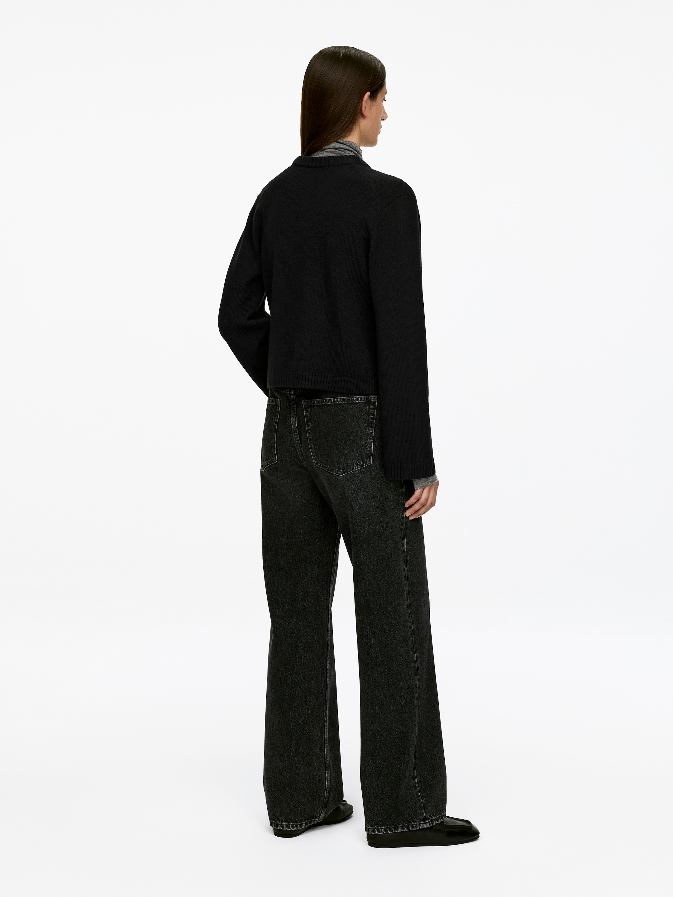 Vis større bilde: En svart crew-neck cardigan med ribbestrikkede mansjetter og en ribbestrikket kant bæres av modellen. Modellen har lagdelt cardiganen over en grå turtleneck.