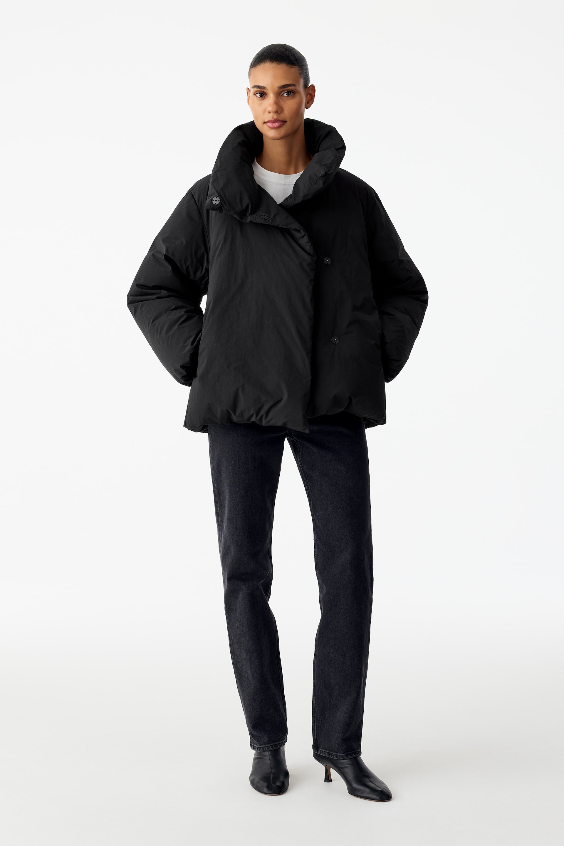 Größeres Bild anzeigen: Eine schwarze Steppjacke mit asymmetrischer Knopfleiste und hohem Stehkragen wird vom Model getragen. Die Jacke wird mit einem weißen T-Shirt im Layering-Look, schwarzen Jeans und schwarzen Stiefeln mit Absatz kombiniert, während das Model die Hände in den Seitentaschen ruht.