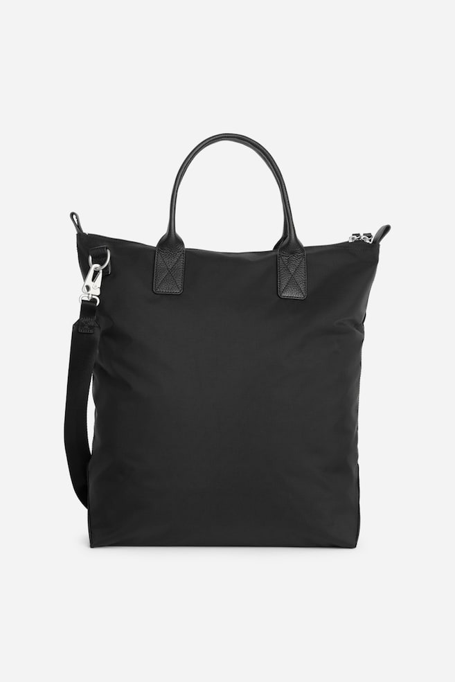 Eine schwarze Nylon-Tote-Bag steht aufrecht in der Frontansicht und verfügt über zwei strukturierte Leder-Tragegriffe oben sowie einen ReiÃverschluss oben. Ein abnehmbarer schwarzer Webbing-Schulterriemen hängt an einem silberfarbenen Metallkarabiner an der Seite. - 1