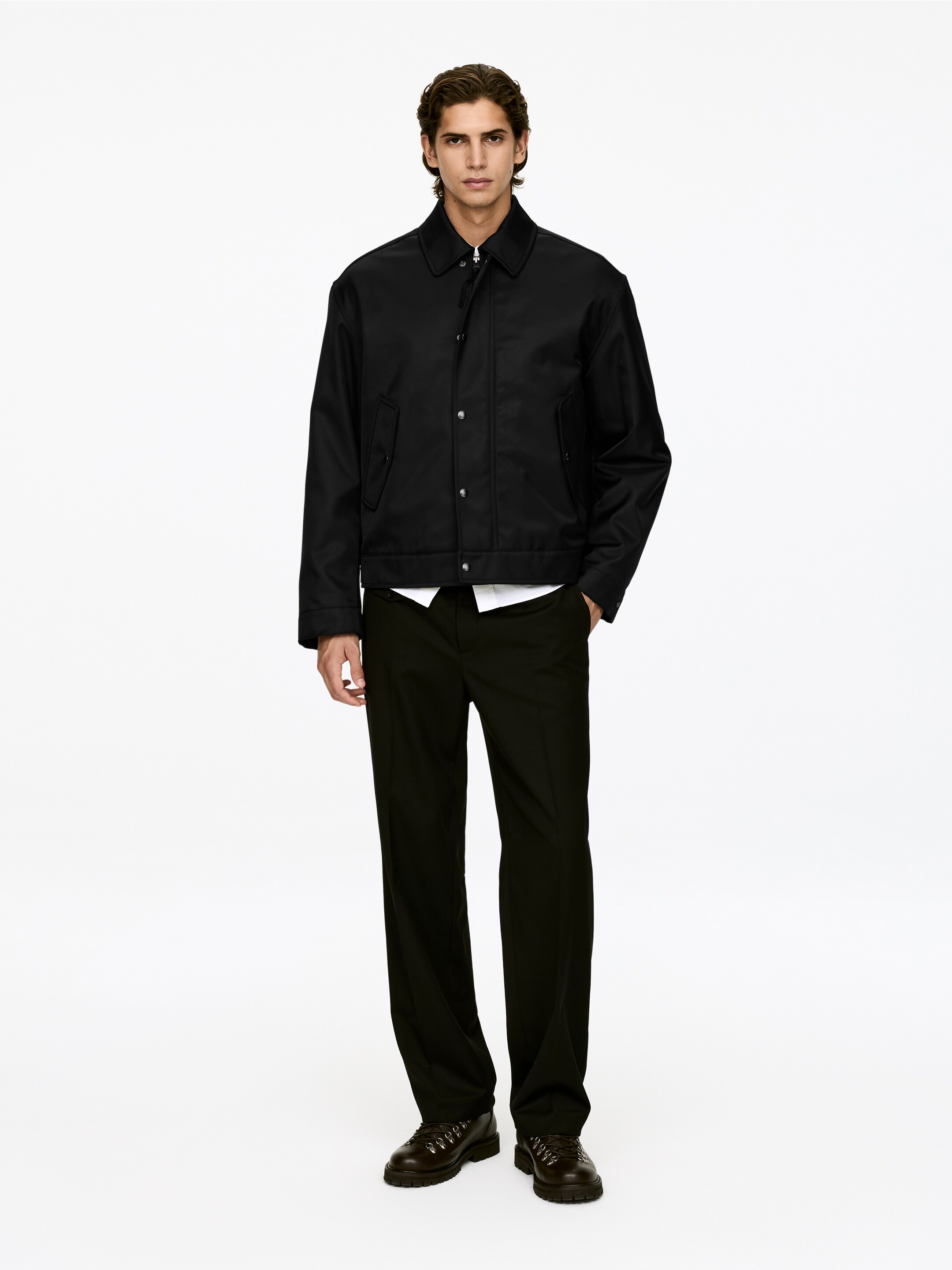 Größeres Bild ansehen: Eine schwarze Jacke mit Kragen wird von dem Model getragen. Sie verfügt über einen vorderen Knopfverschluss mit sichtbaren silbernen Knöpfen und zwei unteren Klappentaschen und ist über einem weißen Hemd gestylt.