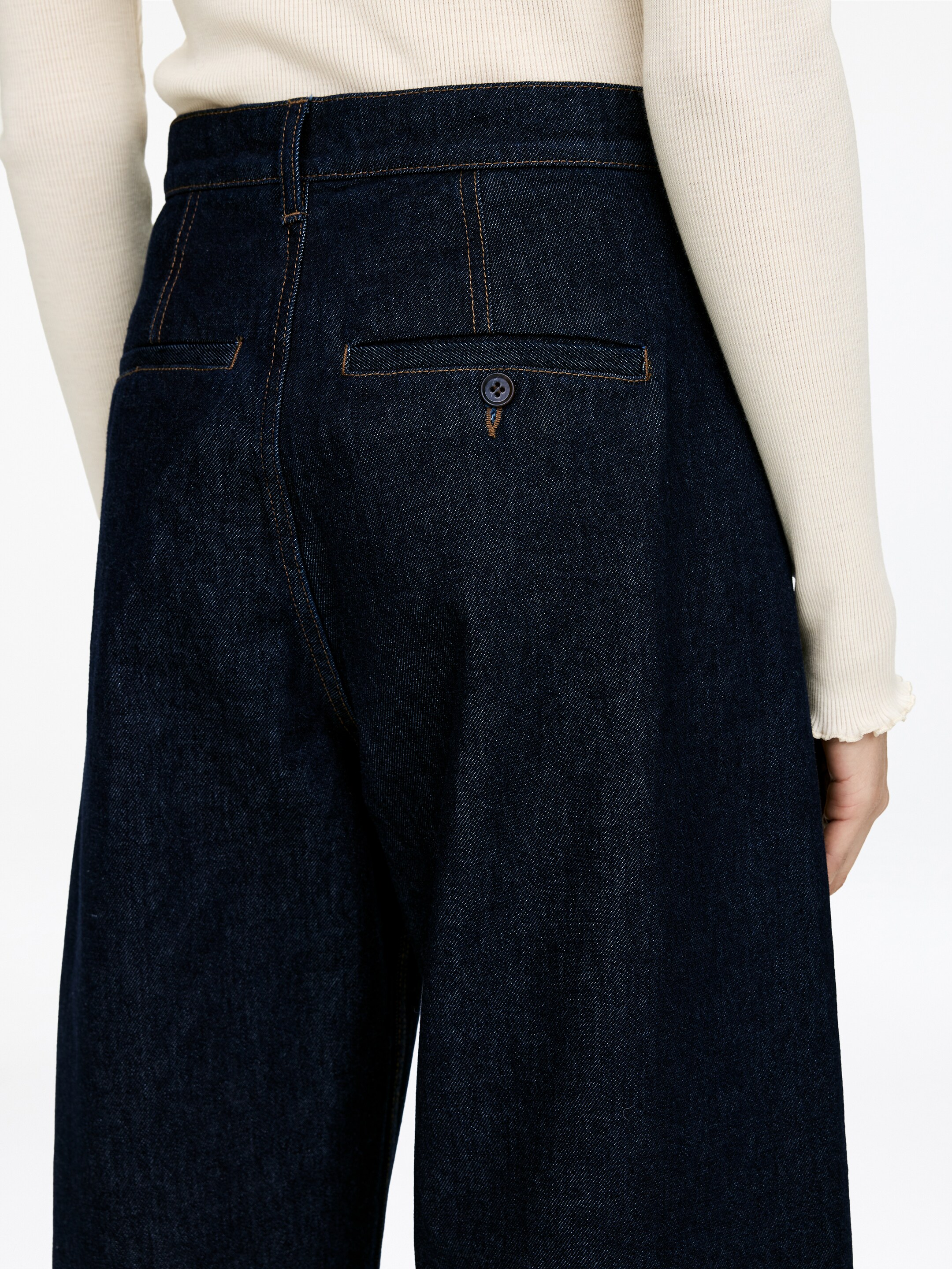 Grotere afbeelding bekijken: Denim jeans in tonvorm met een donkere indigo wassing worden gedragen door het model, met een middenhoge taille en geknoopte paspelzakken aan de achterkant. Contrasterend stiksel accentueert de zakken en naden, en een lichtgekleurde geribde top wordt in de broek gestopt.
