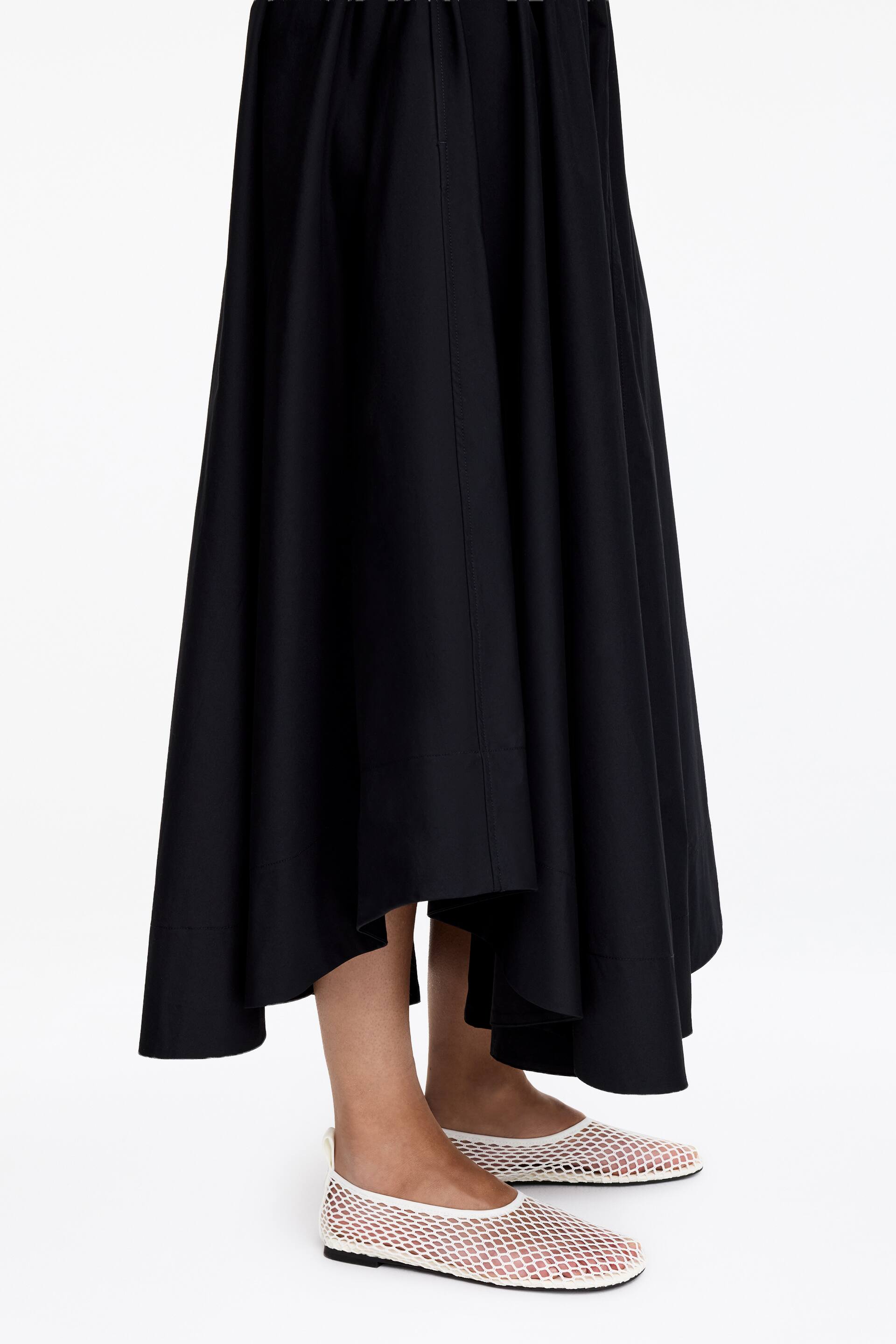 Visualizza immagine più grande: Una gonna lunga nera e fluida è mostrata da un'angolazione laterale, caratterizzata da una vita arricciata e un orlo asimmetrico con un ampio pannello inferiore. È indossata con scarpe piatte in rete bianca.