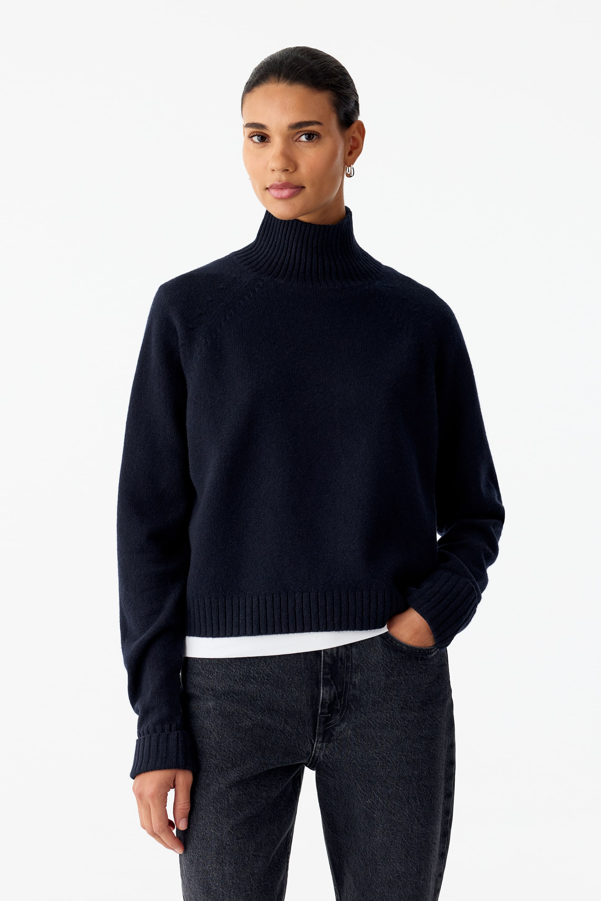 Se større billede: En marineblå strikket rullekravejumper med raglærmer og ribkanter bæres af modellen over en lagdelt hvid T-shirt. Modellen står med den ene hånd hvilende i lommen på mørkegrå jeans.