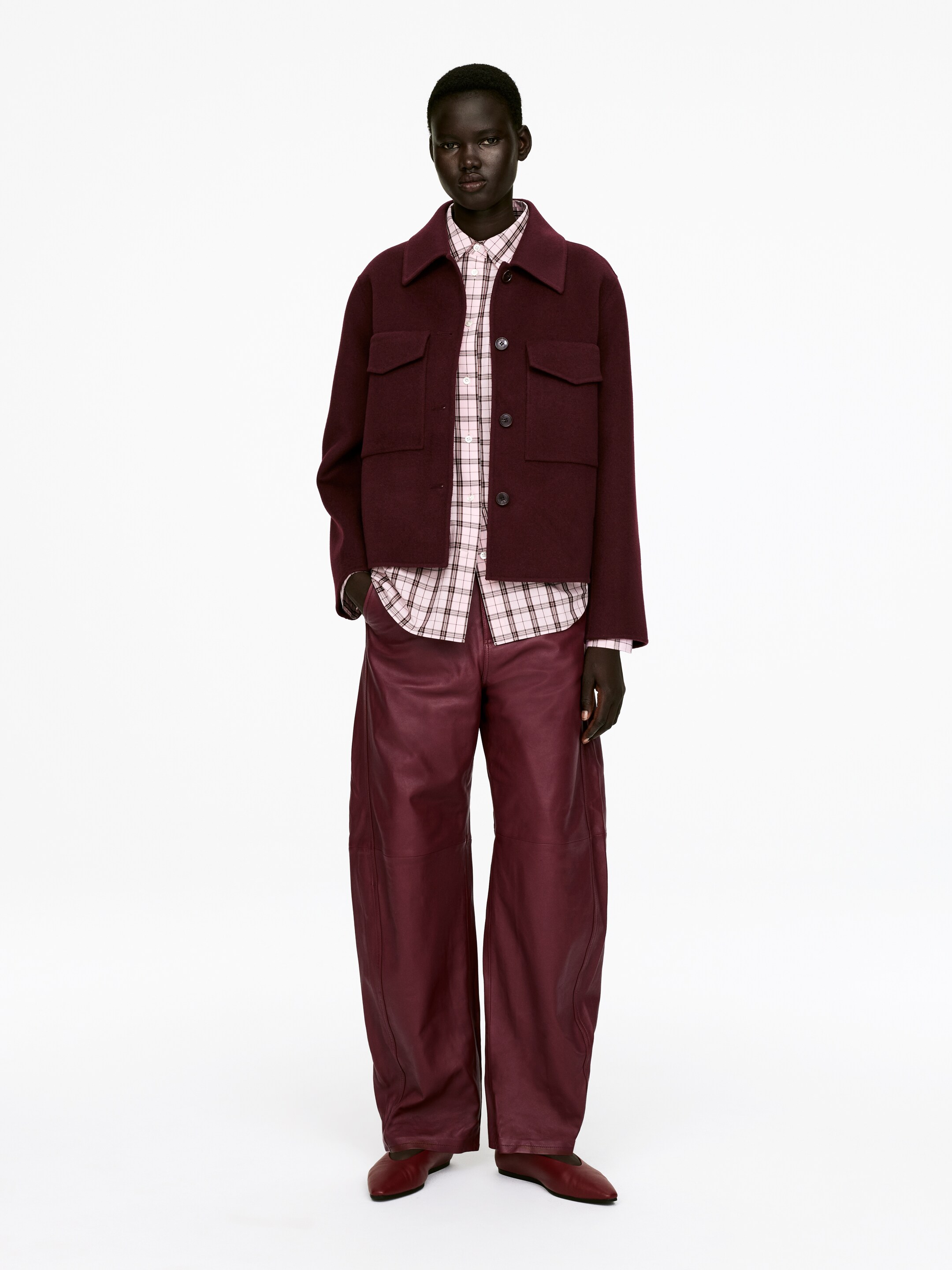 Ver imagen más grande: Una chaqueta de estilo camisa de color rojo oscuro y de corte cuadrado con dos bolsillos con solapa en el pecho se muestra superpuesta sobre una camisa de cuadros rosa claro y blanco, y combinada con pantalones de cuero de pierna ancha a juego en rojo oscuro. Los pantalones presentan líneas de costura visibles y se acumulan en los tobillos, mientras que unos zapatos planos de punta en rojo oscuro completan el conjunto.