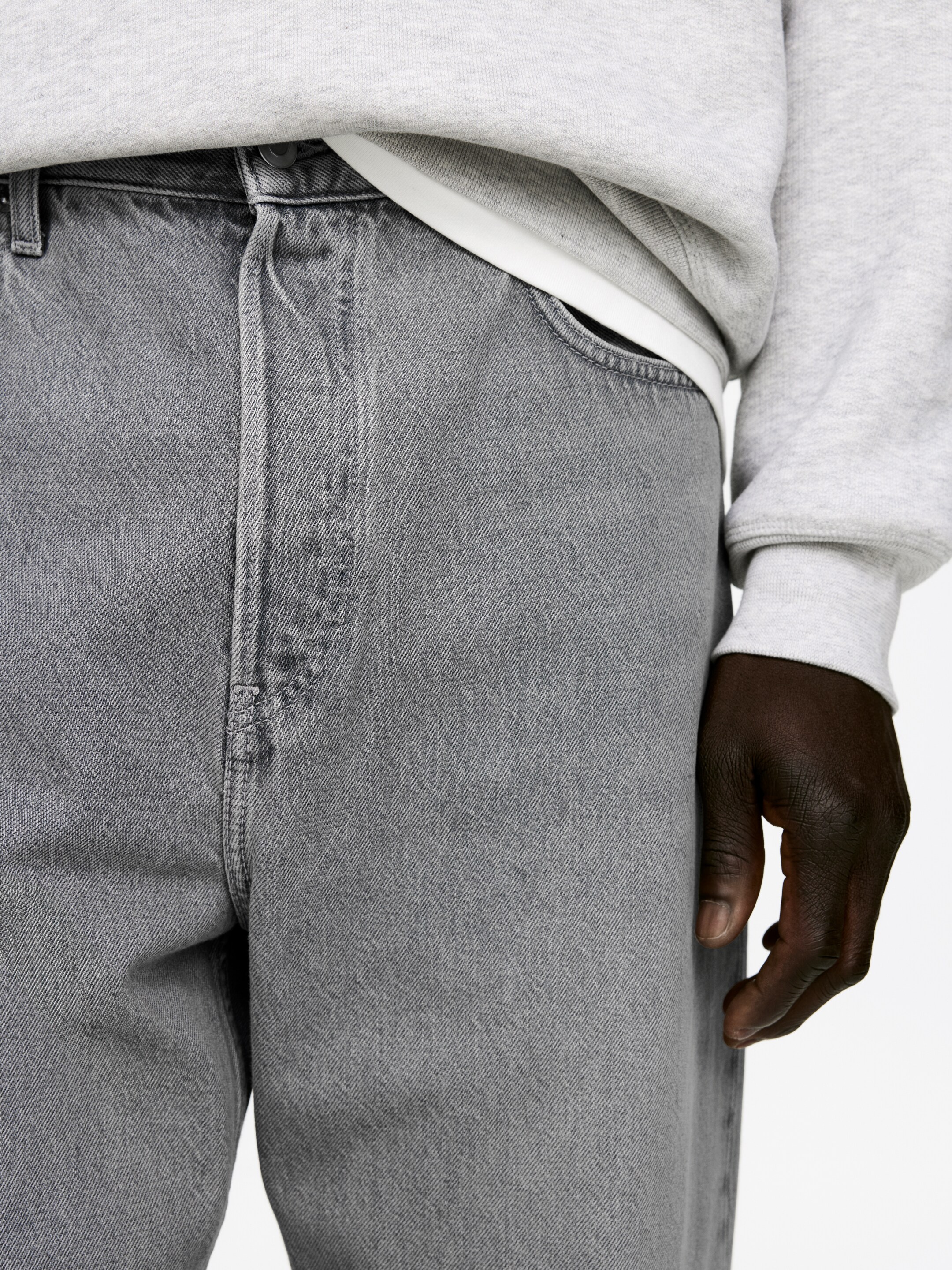 Agrandir l'image: Un jean en denim gris est présenté sous un angle avant-droit, affichant une coupe décontractée et une texture délavée. Le jean présente une ceinture visible avec une fermeture à bouton et une ouverture de poche avant, avec un sweat-shirt gris clair et un sous-vêtement blanc rentré dedans.