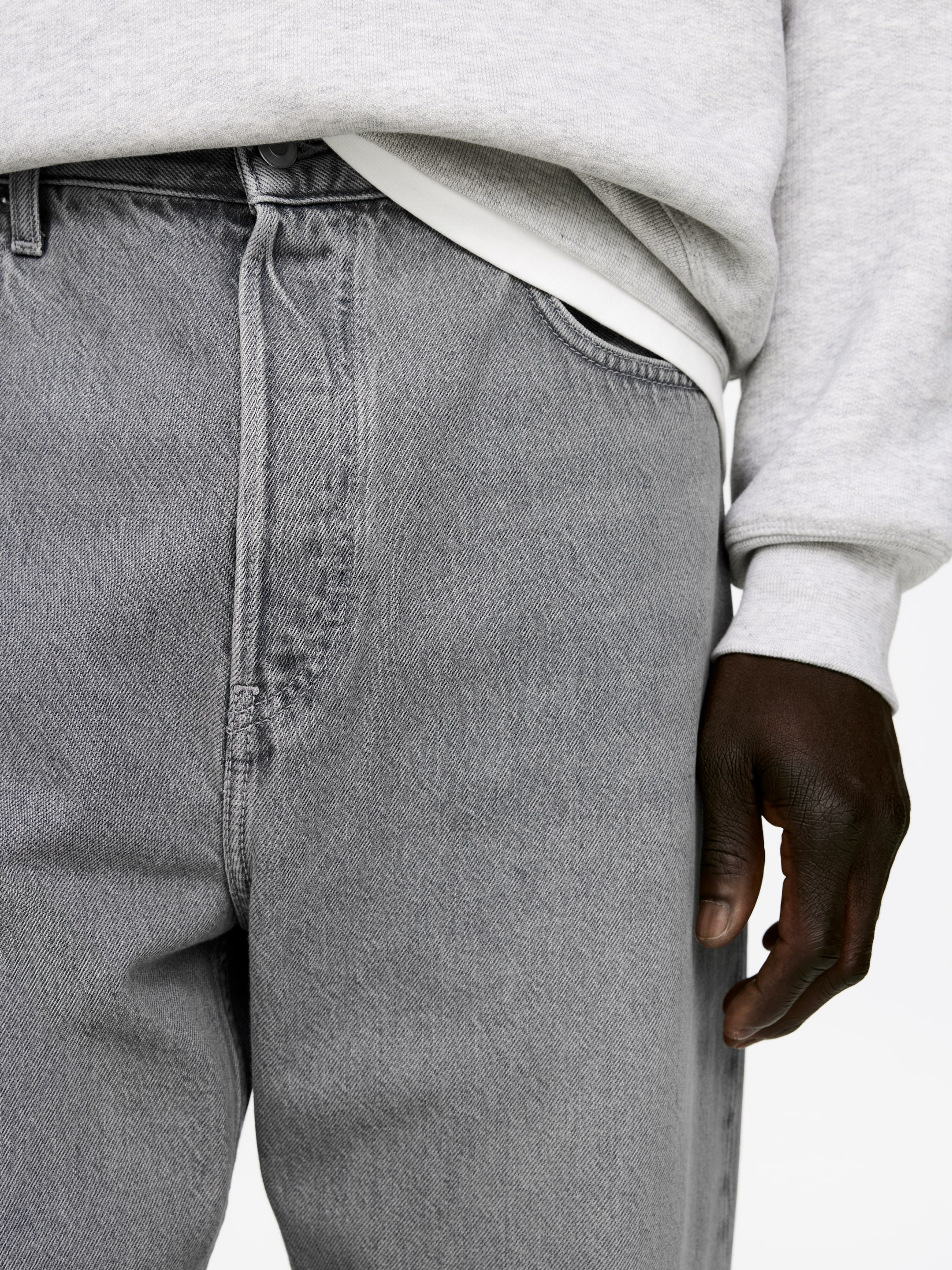 Ingrandisci l'immagine: I jeans in denim grigio sono mostrati da un'angolazione frontale-destra, con una vestibilità rilassata e una texture lavata. I jeans presentano una cintura visibile con chiusura a bottone e un'apertura della tasca anteriore, con una felpa grigio chiaro e una maglietta bianca infilata dentro.