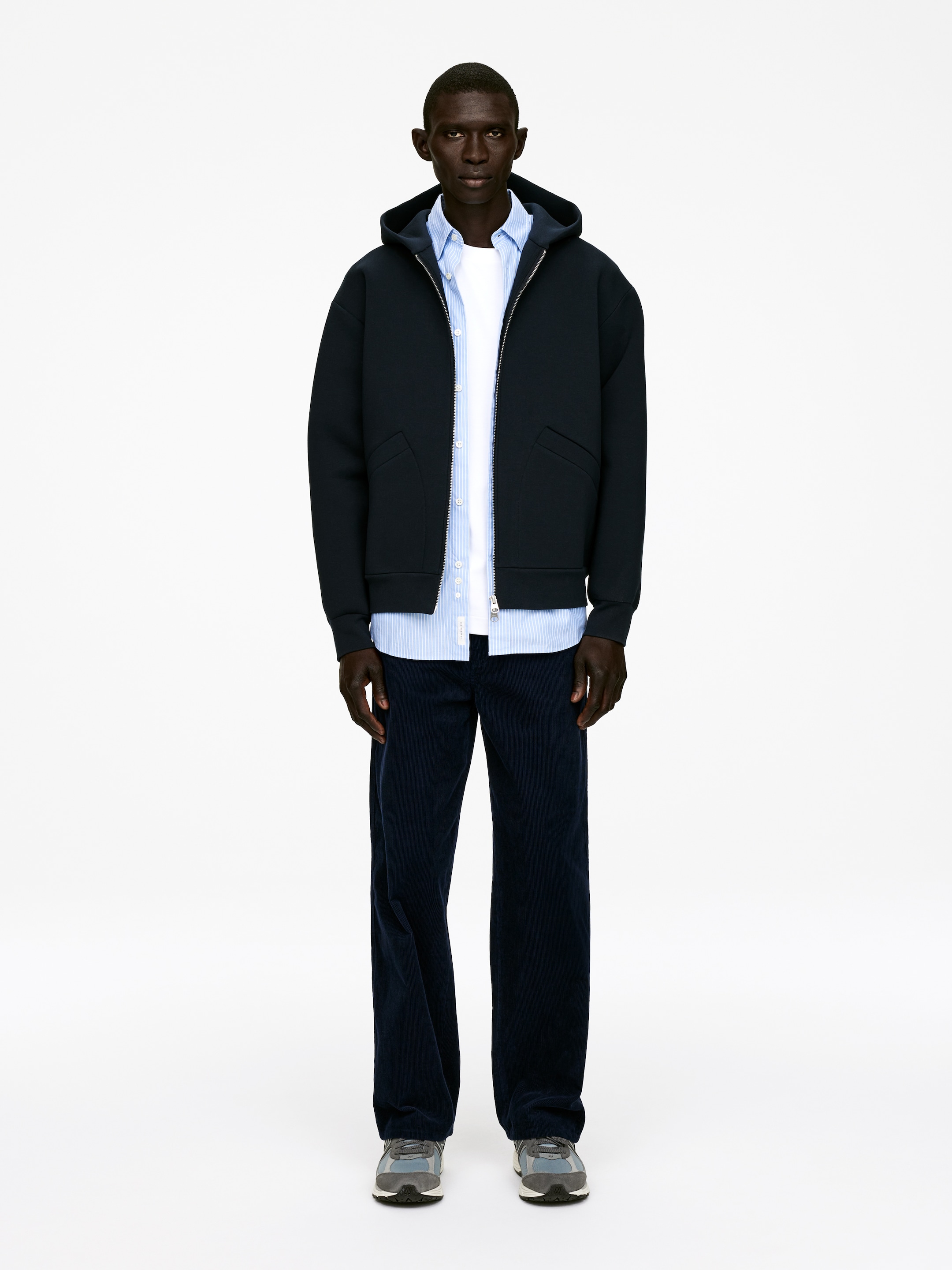 Agrandir l'image: Une veste à capuche bleu foncé est portée ouverte sur un t-shirt blanc et une chemise à col rayée bleu clair. La veste présente une fermeture éclair argentée complète et des poches passepoilées inclinées, et est stylée avec un pantalon en velours côtelé bleu foncé et des chaussures de sport grises et bleues, l'ensemble étant présenté de face.