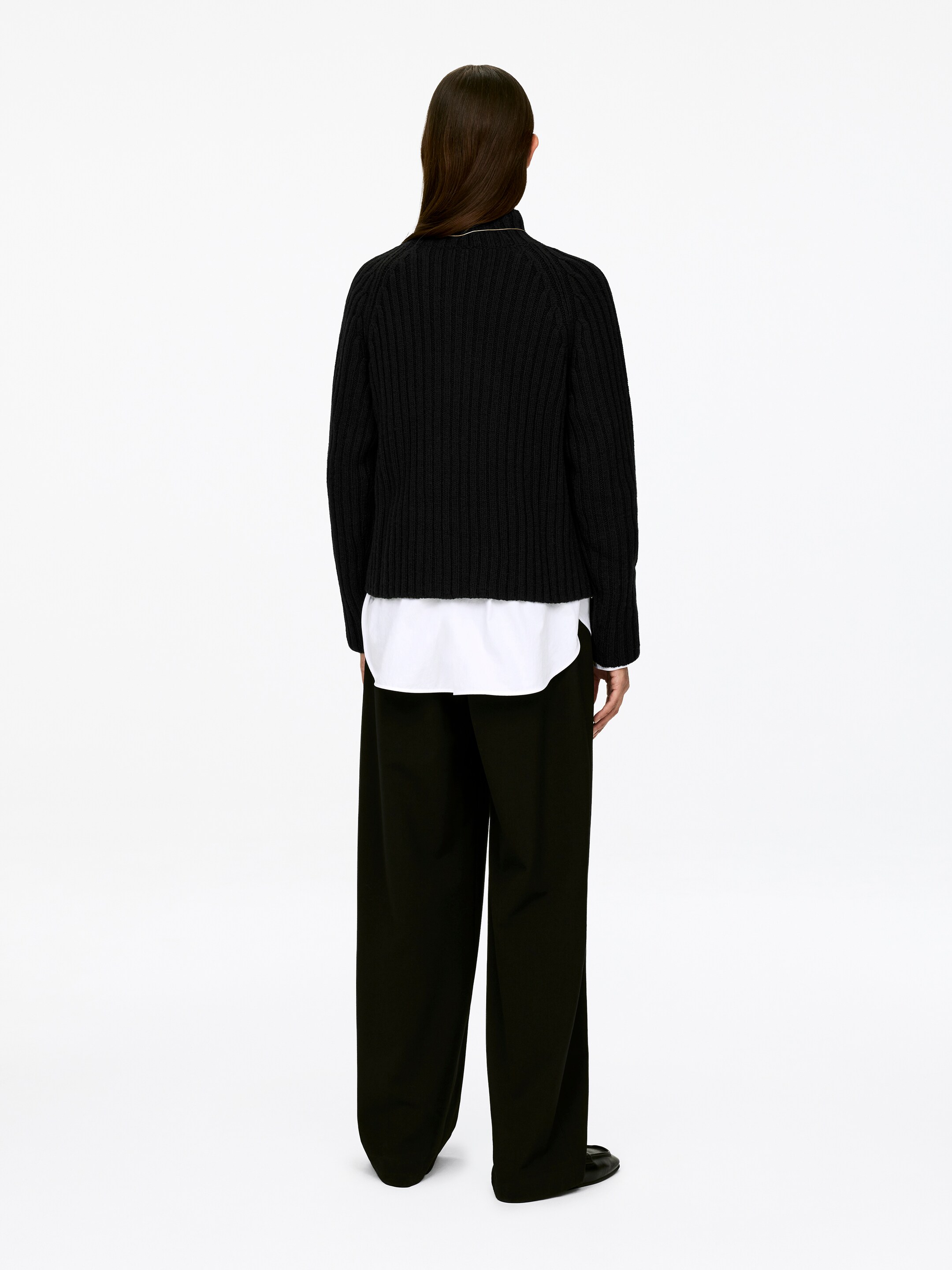 Agrandir l'image: Un pull noir en maille côtelée est porté par le mannequin, avec un col montant et des manches longues. Une chemise blanche est superposée sous le pull, avec son ourlet visible.