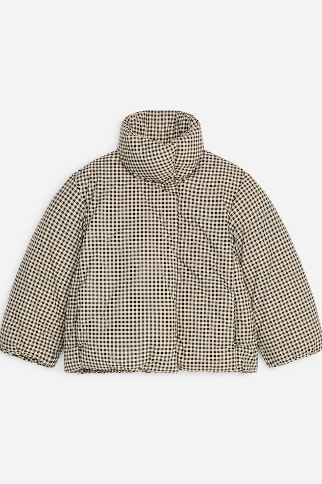 Un piumino con motivo gingham nero e beige è mostrato frontalmente, mettendo in risalto il design imbottito e l'ampio colletto alto a imbuto. La giacca presenta maniche lunghe e ampie e una chiusura frontale nascosta con orlo arricciato. - 1