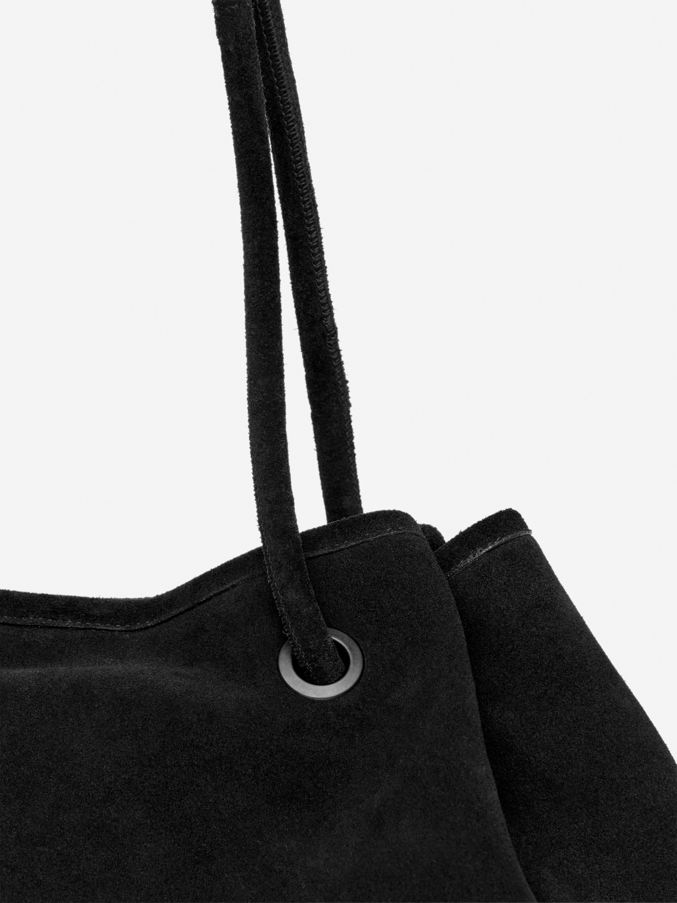 Agrandir l'image: Un sac fourre-tout en daim noir est incliné vers la droite, avec ses bretelles tubulaires glissant à travers des œillets en métal sombre. La texture douce du daim du sac est visible le long de son bord supérieur.