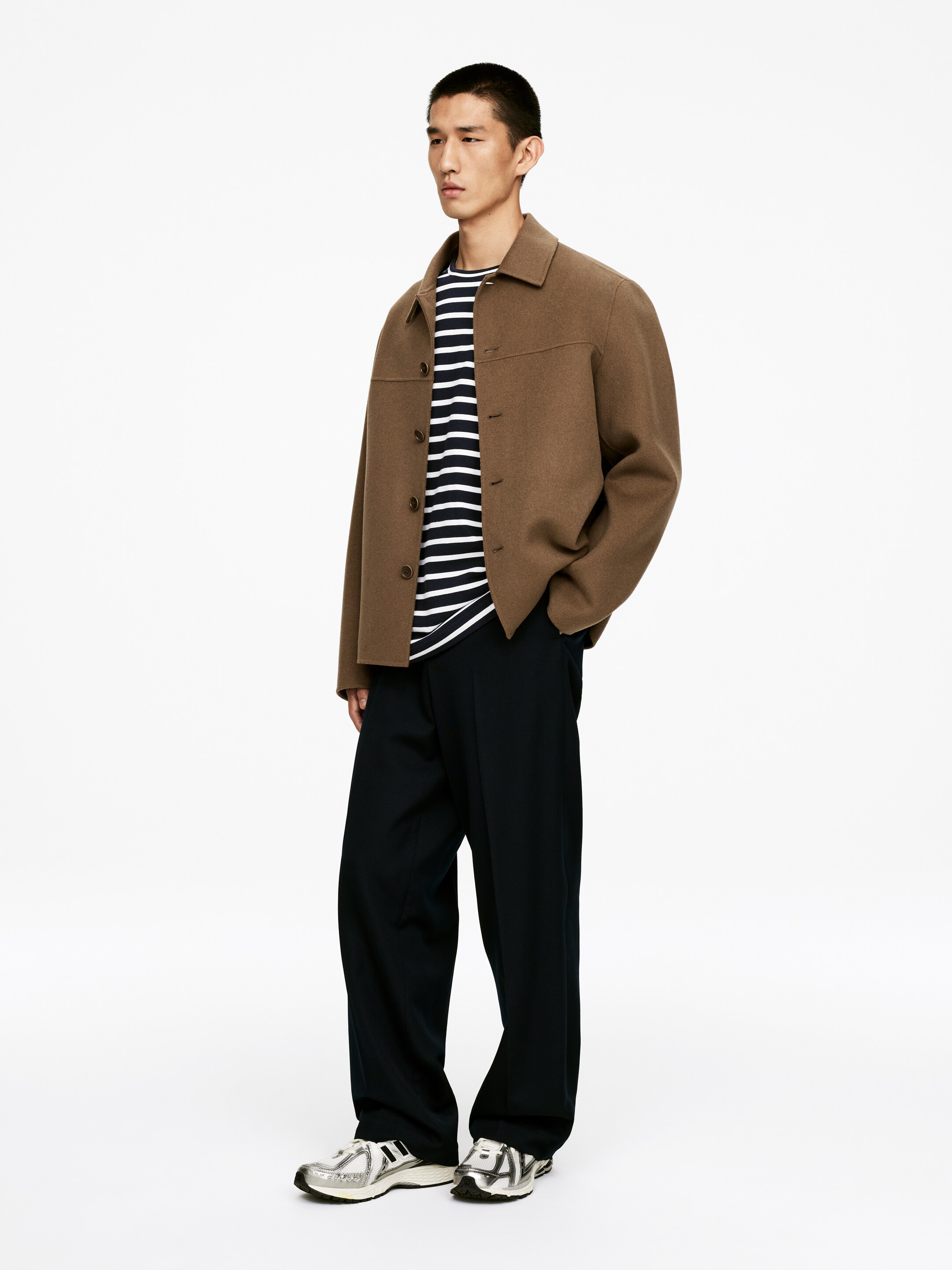 Grotere afbeelding bekijken: Een bruine relaxed-fit button-front jas met een spread kraag wordt gepresenteerd op een model dat iets naar rechts kijkt. Het is gelaagd over een wit en marine gestreept crewneck t-shirt en wordt gecombineerd met zwarte relaxed-fit straight-leg broeken en zilveren en witte chunky sneakers.