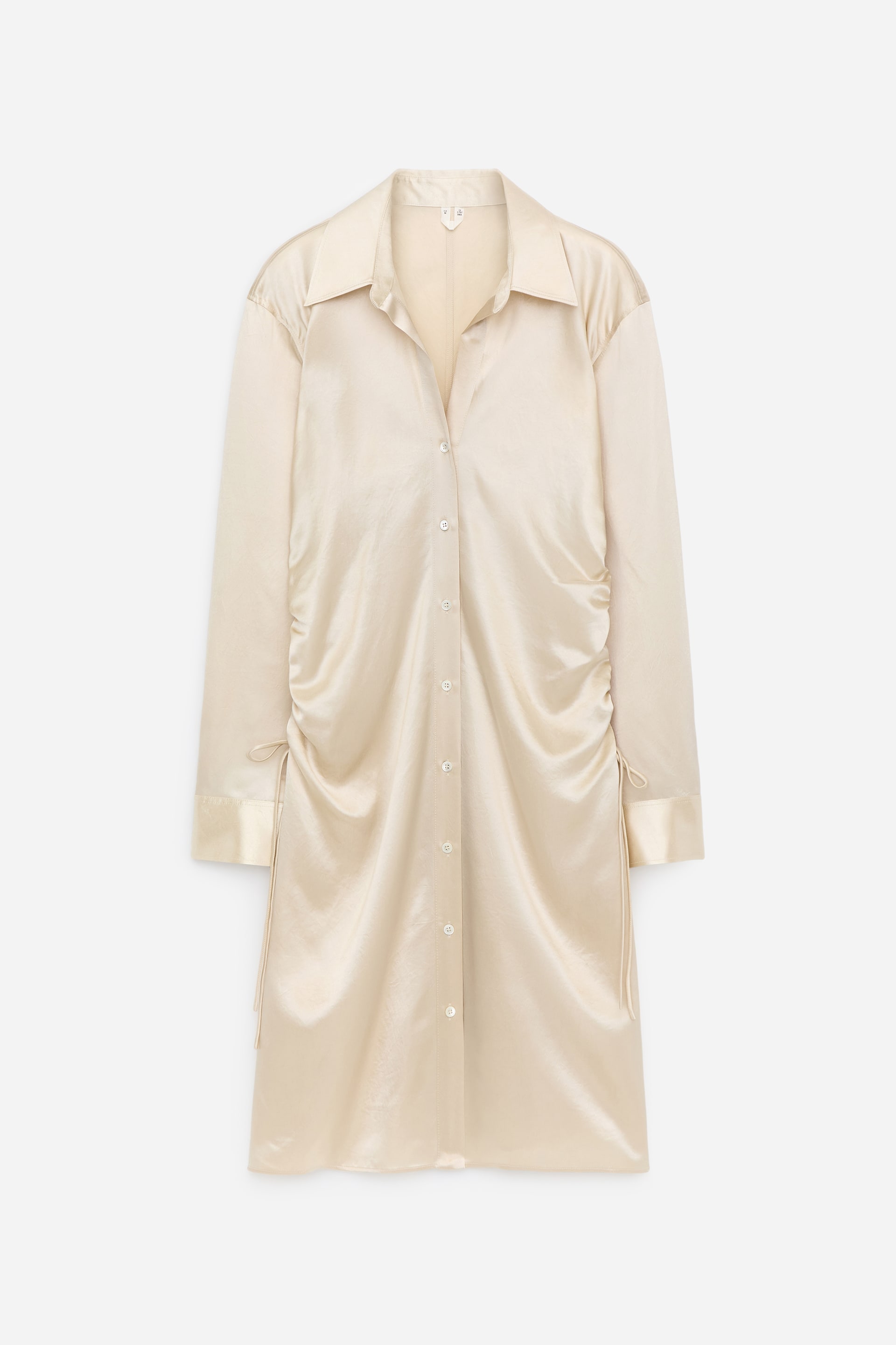 Größeres Bild ansehen: Ein cremefarbenes Satin-Hemdkleid zeigt seine VORDERSEITE, mit einem spitzen Kragen, einer durchgehenden Knopfleiste, langen Ärmeln mit Knopfmanschetten und einer sanften seitlichen Raffung, geformt durch zarte Kordelzüge.