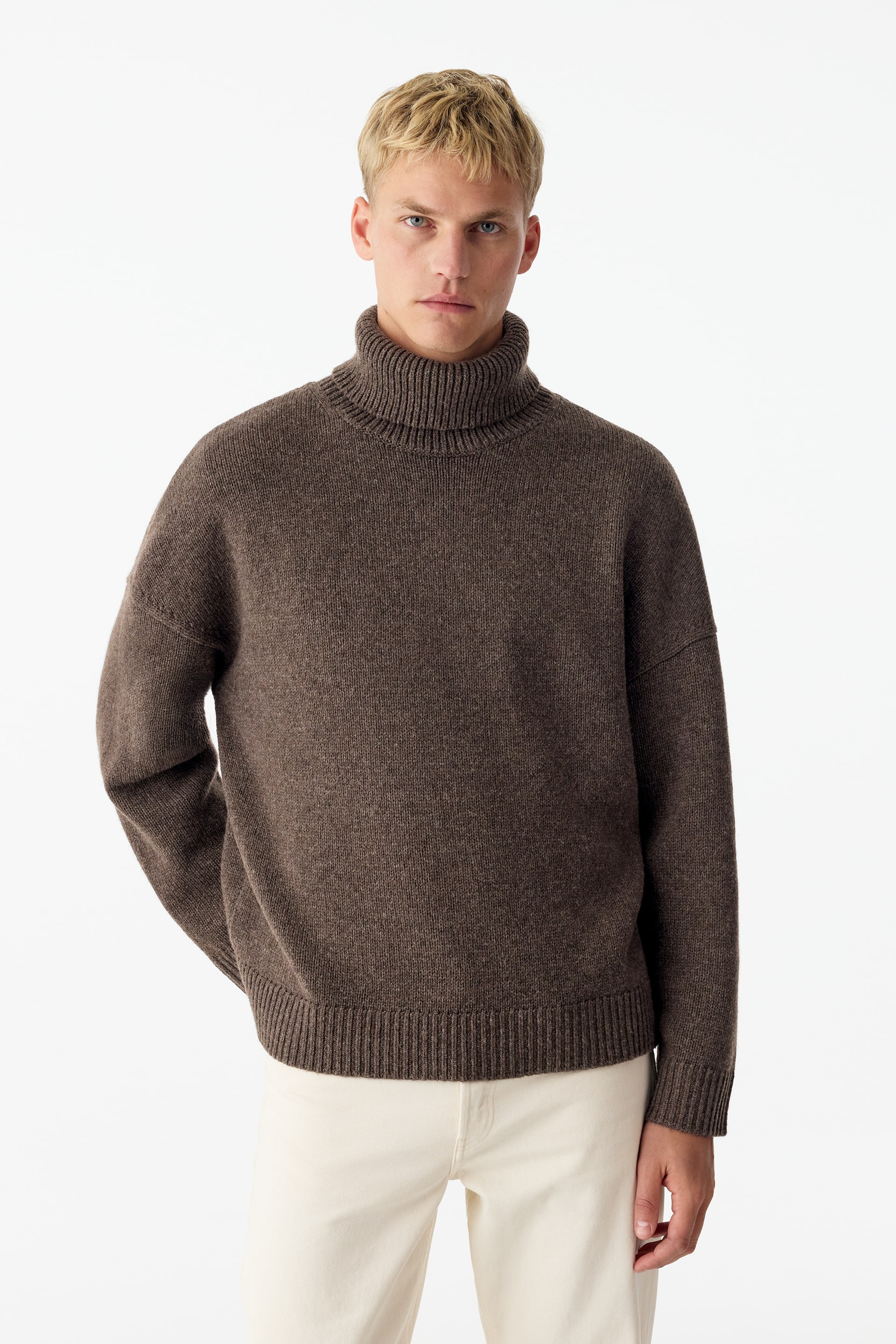 View larger image: A brown, relaxed-fit roll-neck jumper made of textured wool is being worn by the model, featuring a high ribbed collar, dropped shoulders, and ribbed cuffs and hem. The jumper is paired with off-white trousers.