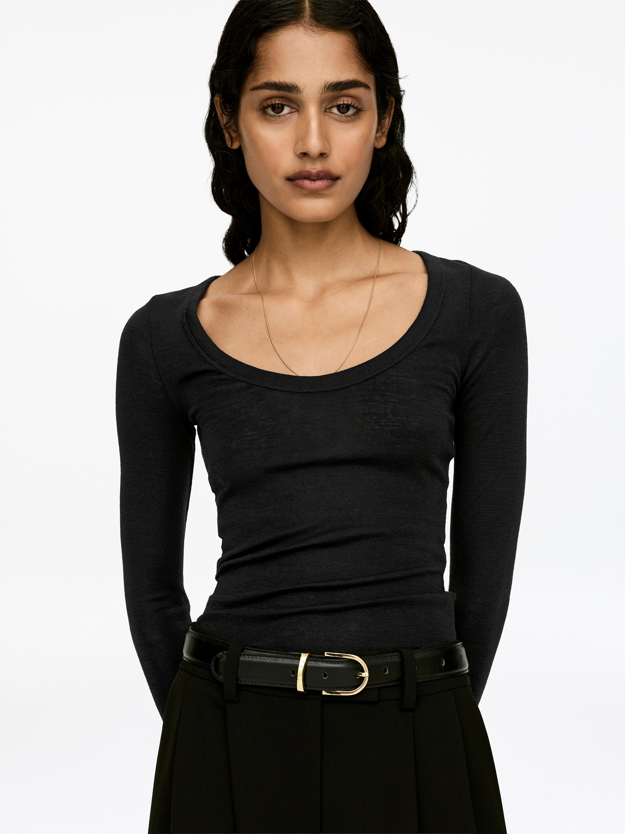 Agrandir l'image: Un haut en laine sombre, tricoté en côtes, est porté par le mannequin, avec une coupe ajustée, des manches longues et un col échancré. Le haut est complété par un pantalon noir et une ceinture noire avec une boucle dorée, et un collier fin en chaîne dorée repose sur le col.
