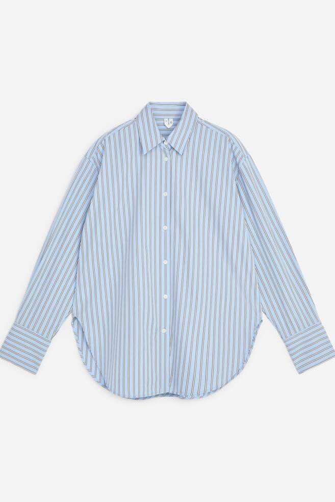 A long-sleeved, button-up shirt is displayed from the front in a light blue with a thin vertical stripe pattern. The shirt features a relaxed silhouette with dropped shoulders, a classic point collar, wide cuffs, and a curved hemline. - 1