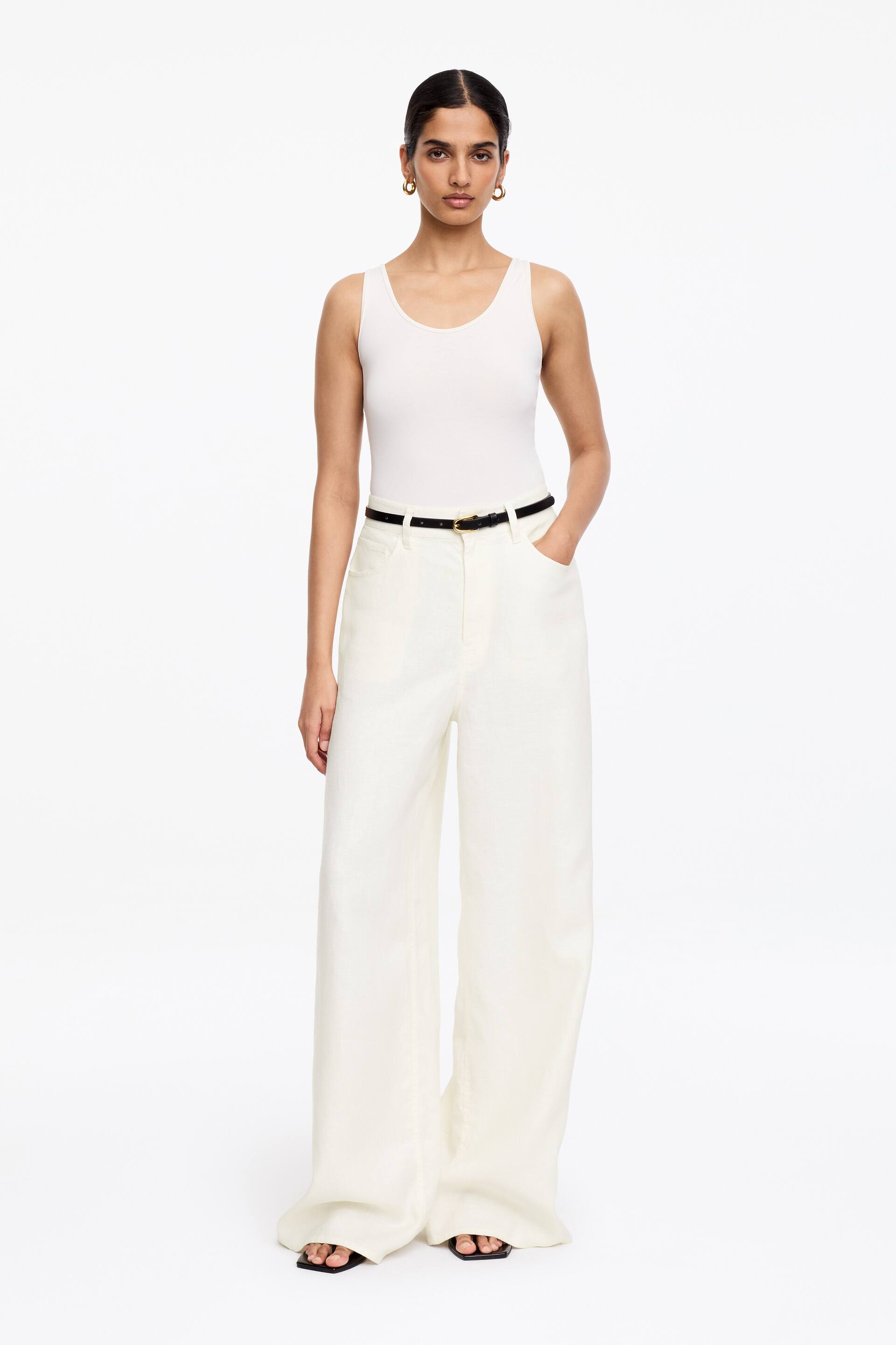Visa större bild: En vit tanktopp med skopringning bärs tillsammans med högmidjade, vida benbyxor i offwhite, som har framfickor och är säkrade i midjan med ett tunt svart bälte med ett guldfärgat spänne.