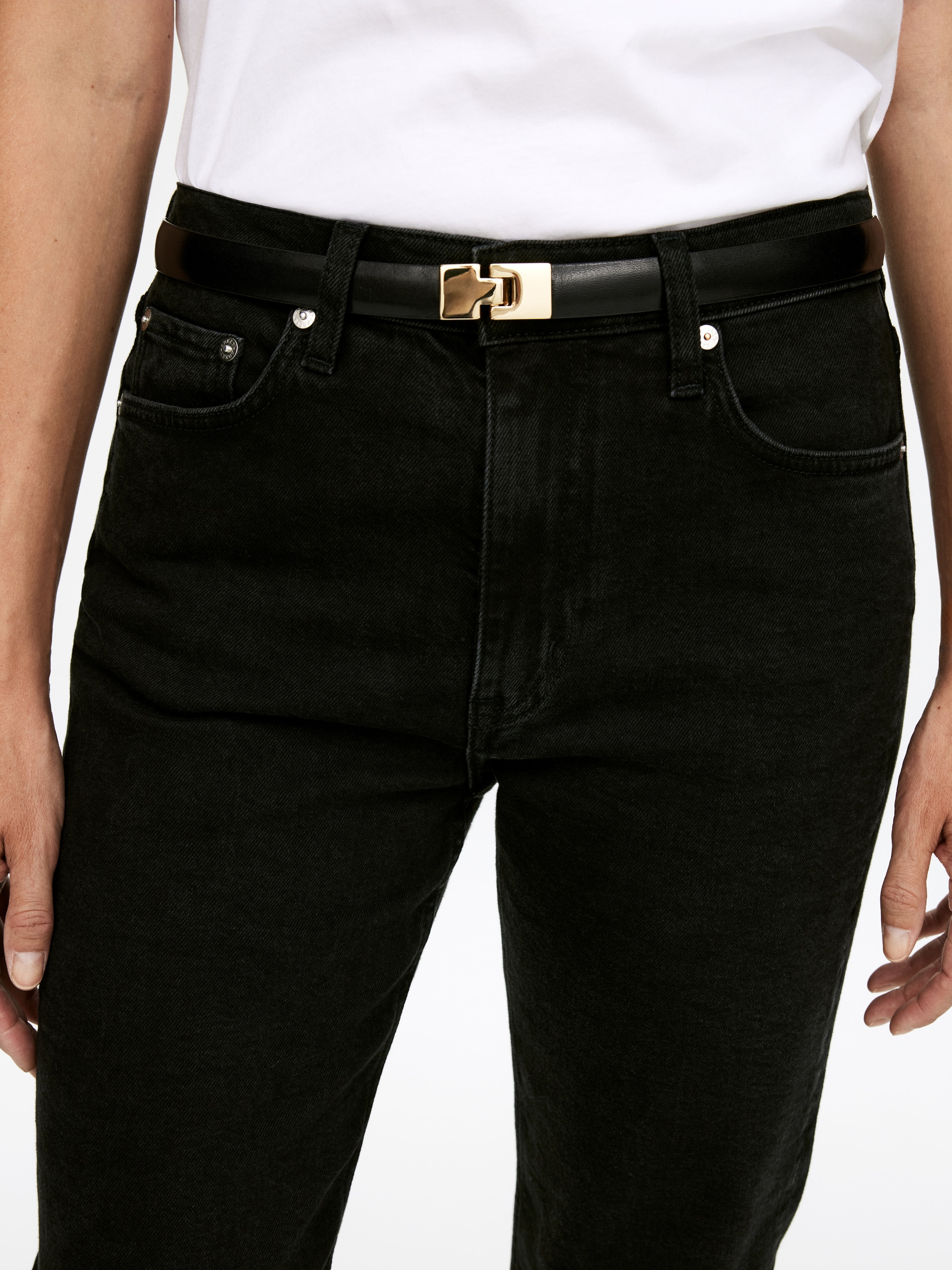 Größeres Bild anzeigen: Ein schmaler schwarzer Gürtel mit einer rechteckigen, goldfarbenen Schnalle wird in der Taille getragen und durch die Gürtelschlaufen einer schwarzen Jeans gefädelt. Ein weißes Oberteil wird in die Jeans gesteckt, wodurch die Platzierung des Gürtels hervorgehoben wird.
