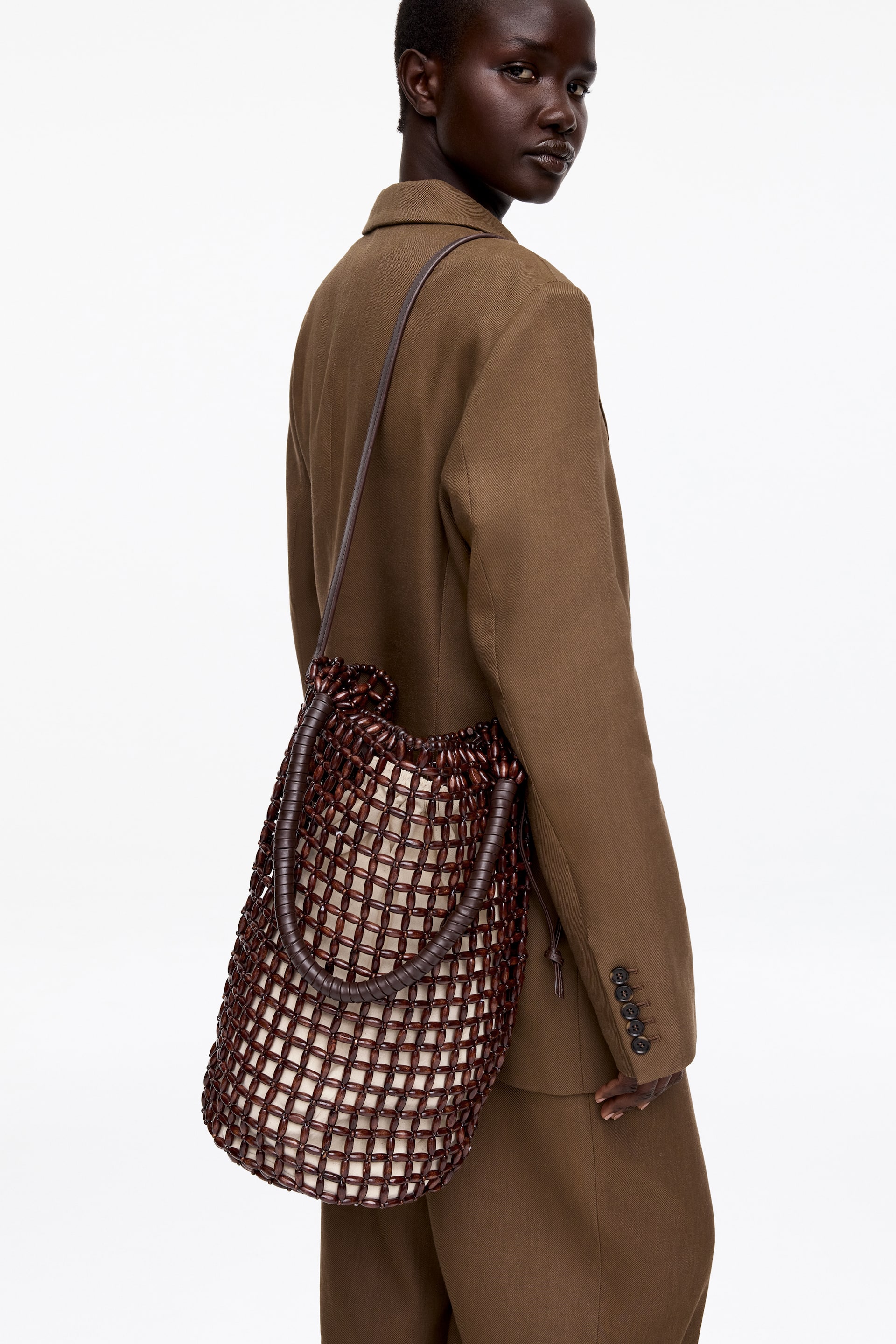 Agrandir l'image: Un sac seau en perles brun foncé est suspendu à une épaule, avec une coque extérieure à motif quadrillé de perles brun foncé sur une doublure intérieure plus claire, et doté de deux poignées arrondies sur le dessus avec une fine bandoulière.