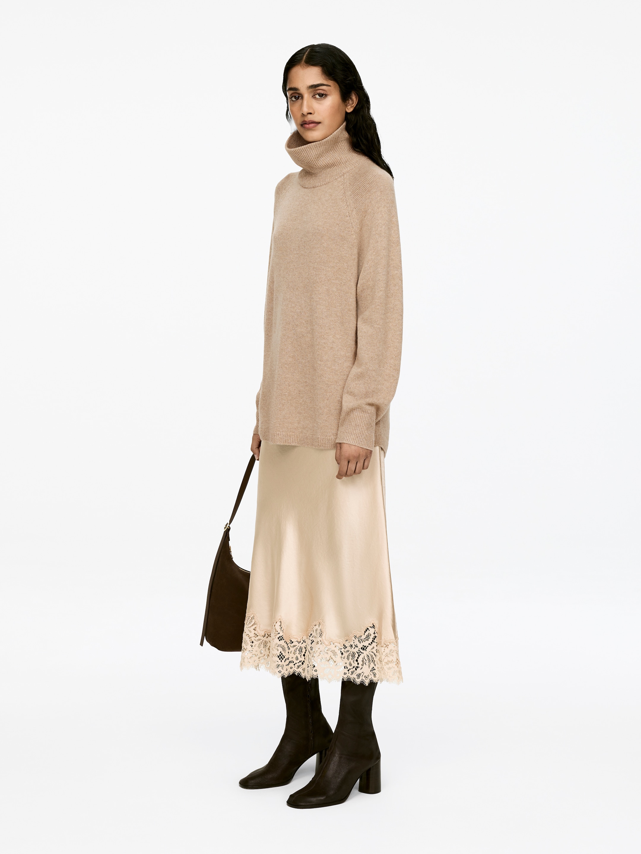 Visa större bild: En beige polotröja med detaljer som raglanärmar och ribbade kanter bärs av modellen. Den höga polokragen täcker modellens hals, och tröjan lagerar över en ljusfärgad slipkjol.