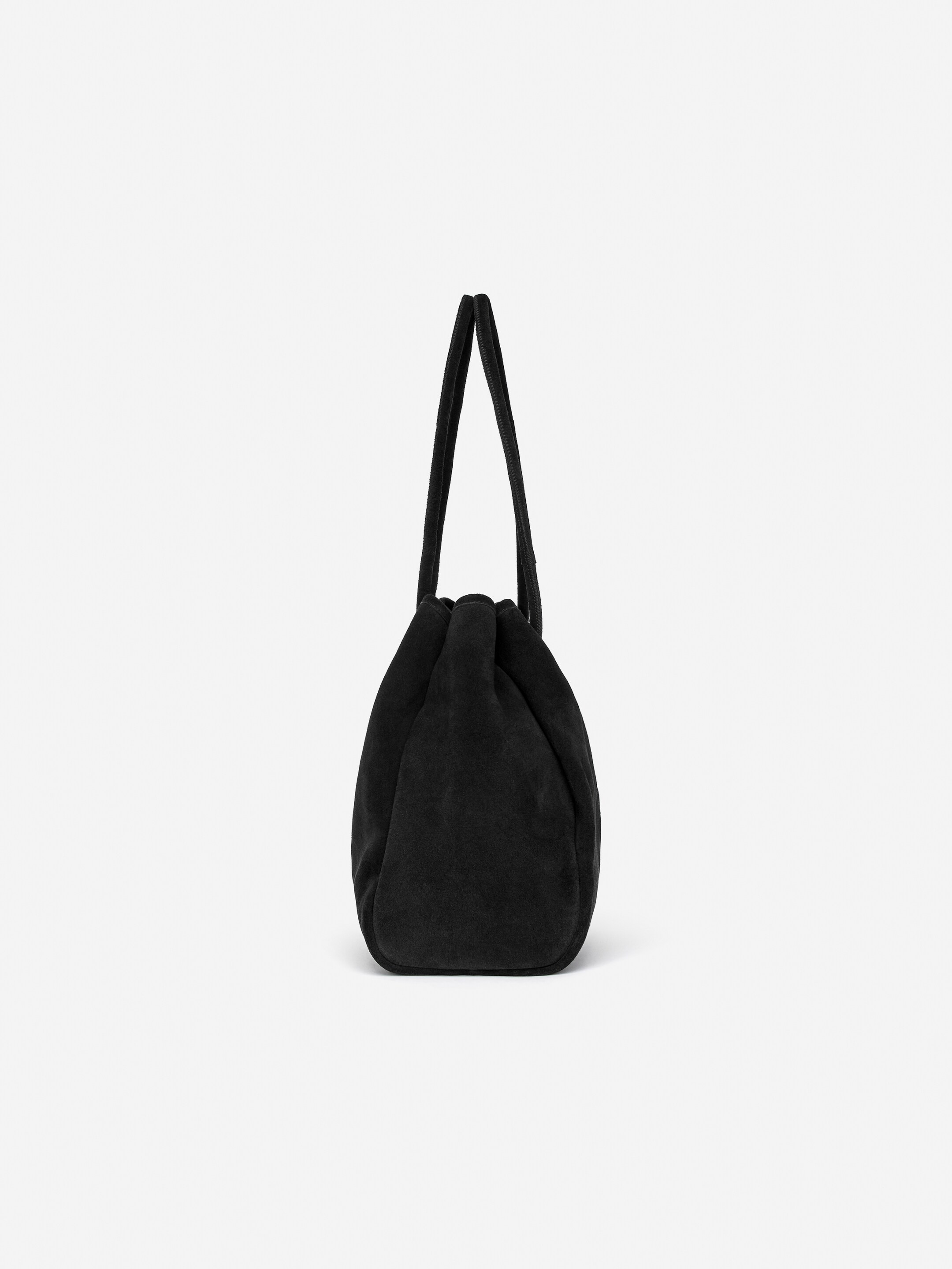 Agrandir l'image: Un sac fourre-tout en daim noir est présenté, son corps doux et arrondi est resserré en haut. Des sangles tubulaires forment les poignées d'épaule, qui ferment également l'ouverture du sac.
