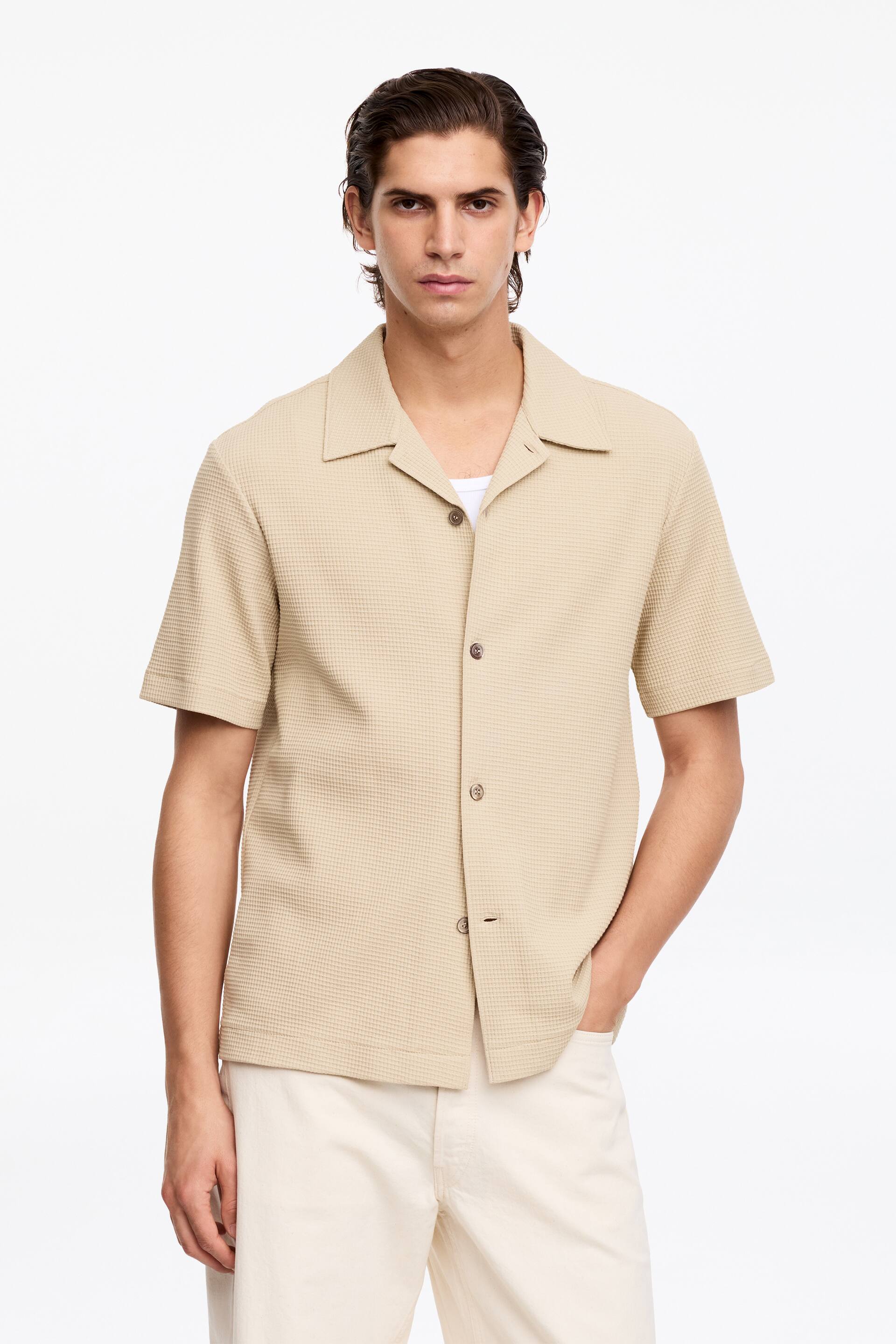 Grösseres Bild ansehen: Ein beiges Kurzarmhemd zeigt eine Waffelstrickstruktur und einen Reverskragen. Es ist mit einer offenen Front gestaltet, wobei drei sichtbare dunkelbraune Knöpfe zu sehen sind, und ein weißes Unterhemd blitzt unter dem offenen Kragen hervor.