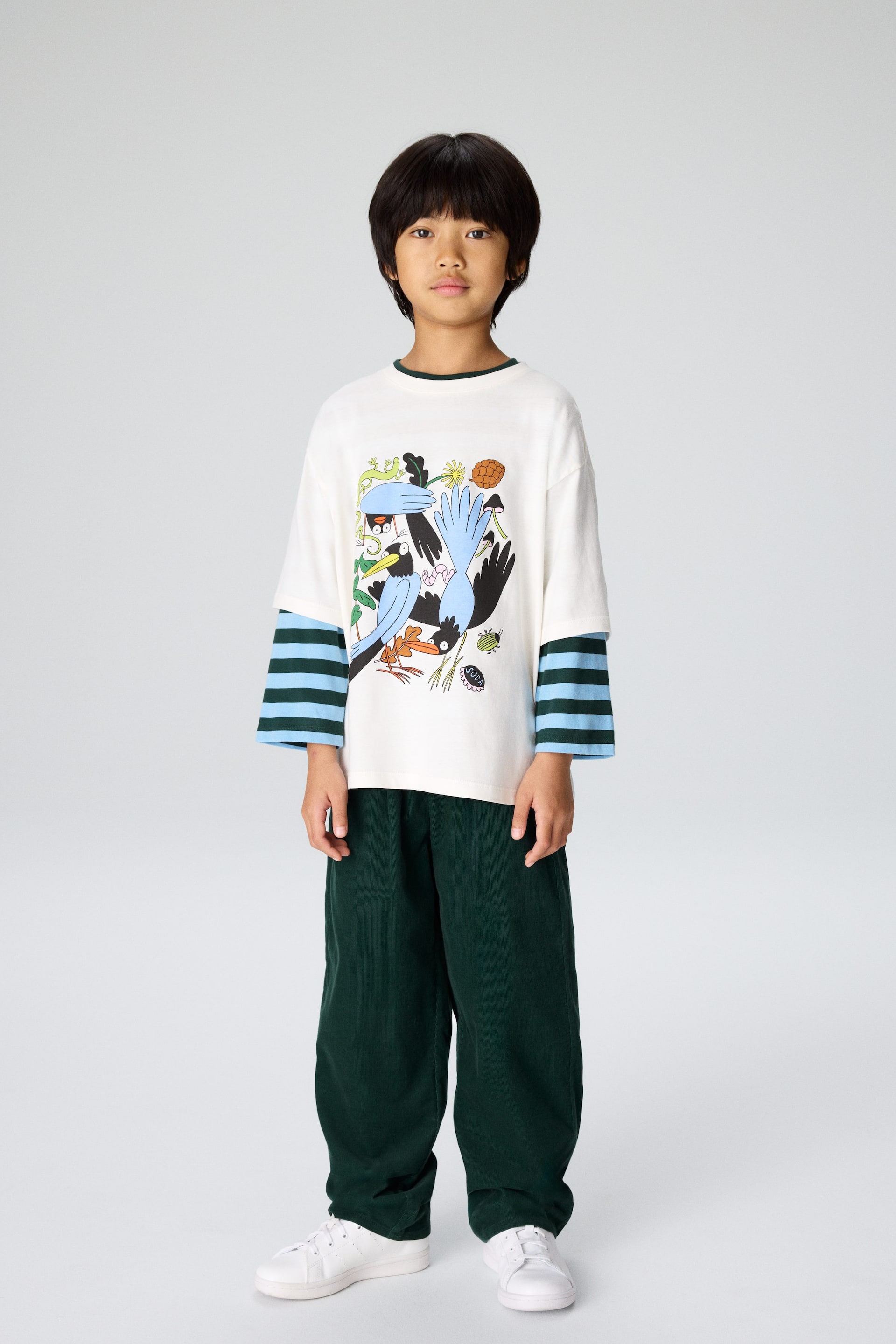 Agrandir l'image: Un t-shirt à manches longues surdimensionné de couleur crème, orné d'un imprimé graphique coloré représentant des oiseaux et des mains stylisés, est superposé à une chemise à manches longues rayée bleu et vert foncé. La tenue vue de face est complétée par un pantalon en velours côtelé vert foncé à jambes larges et des baskets blanches.