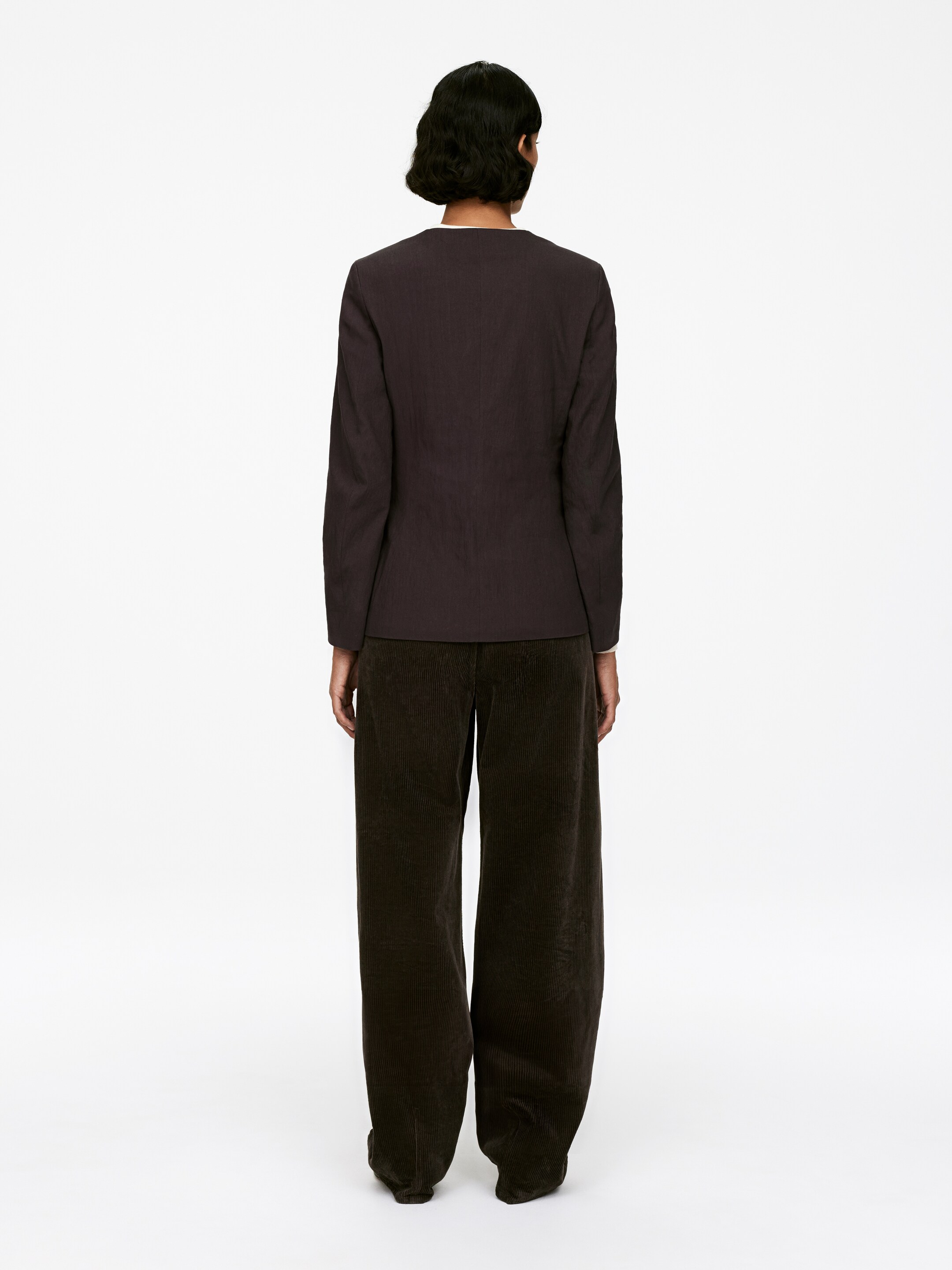 Visualizza immagine più grande: Un blazer scuro, senza colletto, tagliato per una vestibilità slim, è indossato dal modello, che sta in piedi con la schiena rivolta verso l'osservatore. Il blazer è abbinato a pantaloni di velluto a coste scuri e a gamba larga.