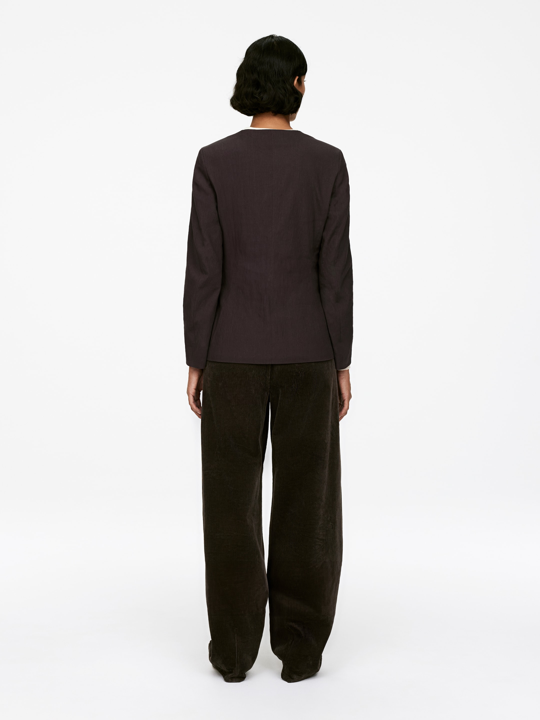 View larger image: A dark, collarless blazer, cut for a slim fit, is being worn by the model, who is standing with their back to the viewer. The blazer is being styled with dark, wide-leg corduroy pants.