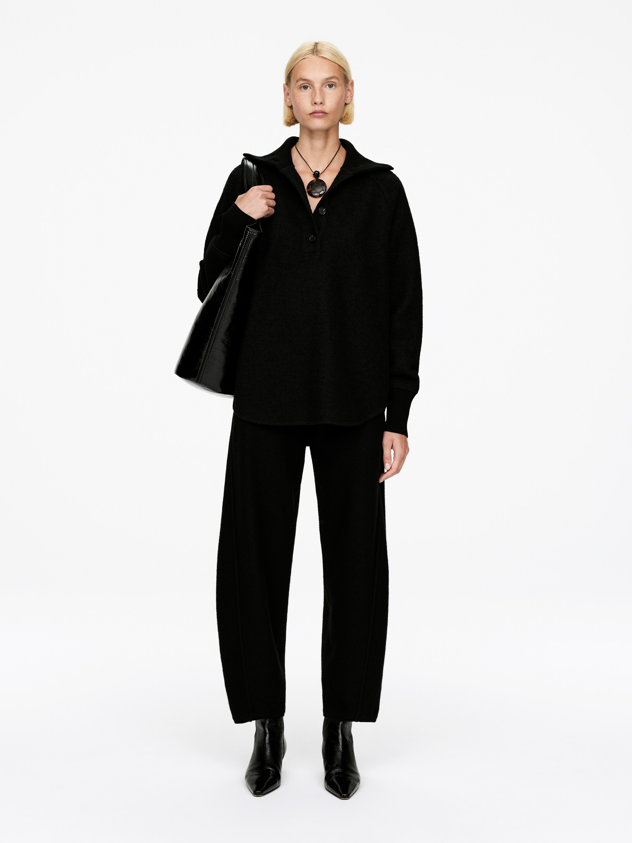 View larger image: A front-facing black polo sweater is being presented, featuring a collared neckline, a three-button placket, and raglan sleeves with ribbed cuffs. It is being styled with black wide-leg pants displaying a visible front seam, and black pointed-toe ankle boots. A black patent tote bag is being held, and a large black circular pendant necklace is being worn over the sweater.