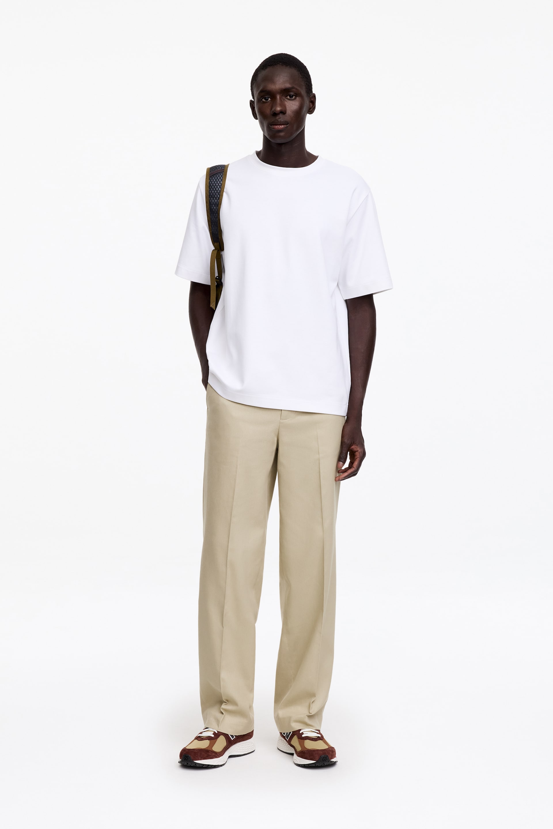 Größeres Bild ansehen: Das Model trägt beigefarbene Wide-Fit-Hosen mit geradem Bein und sichtbarem Bund, wobei eine Hand in einer Seitentasche steckt. Die Hose wird mit einem weißen T-Shirt und Sneakers präsentiert.
