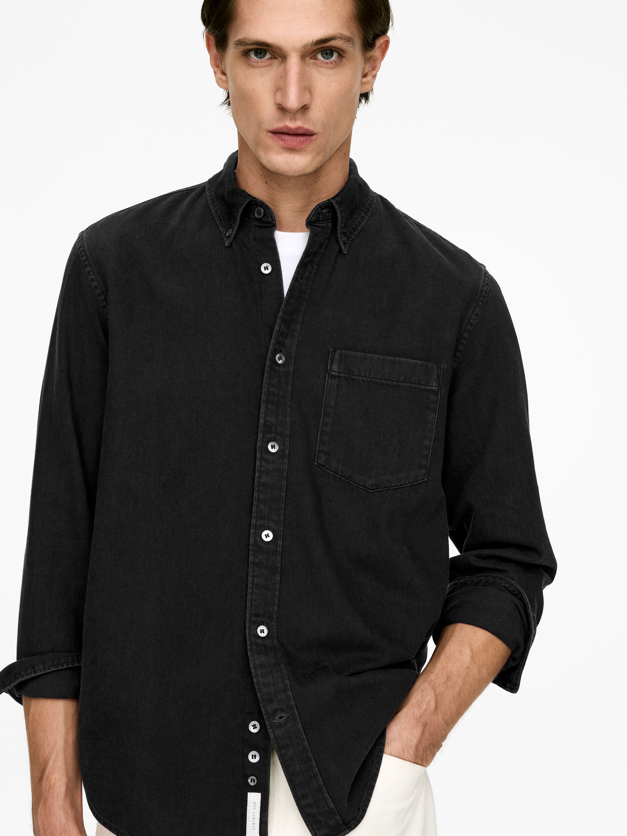 Agrandir l'image: Une chemise noire, texturée, est présentée de face, avec un col boutonné, une poche plaquée unique sur la poitrine, et des boutons blancs le long de la patte de boutonnage. Ses manches longues sont retroussées, et elle est portée par-dessus un t-shirt blanc.