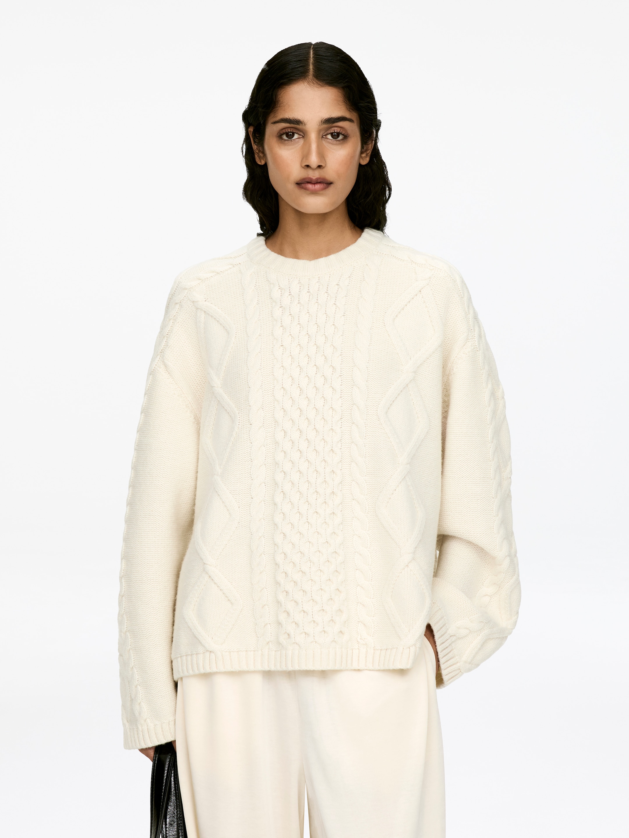 View larger image: A cream wool cable knit jumper with a crew neck, long sleeves, and ribbed trims is being worn by the model. The relaxed-fit jumper features prominent cable patterns, and the model is placing one hand into a pocket of light-colored pants.