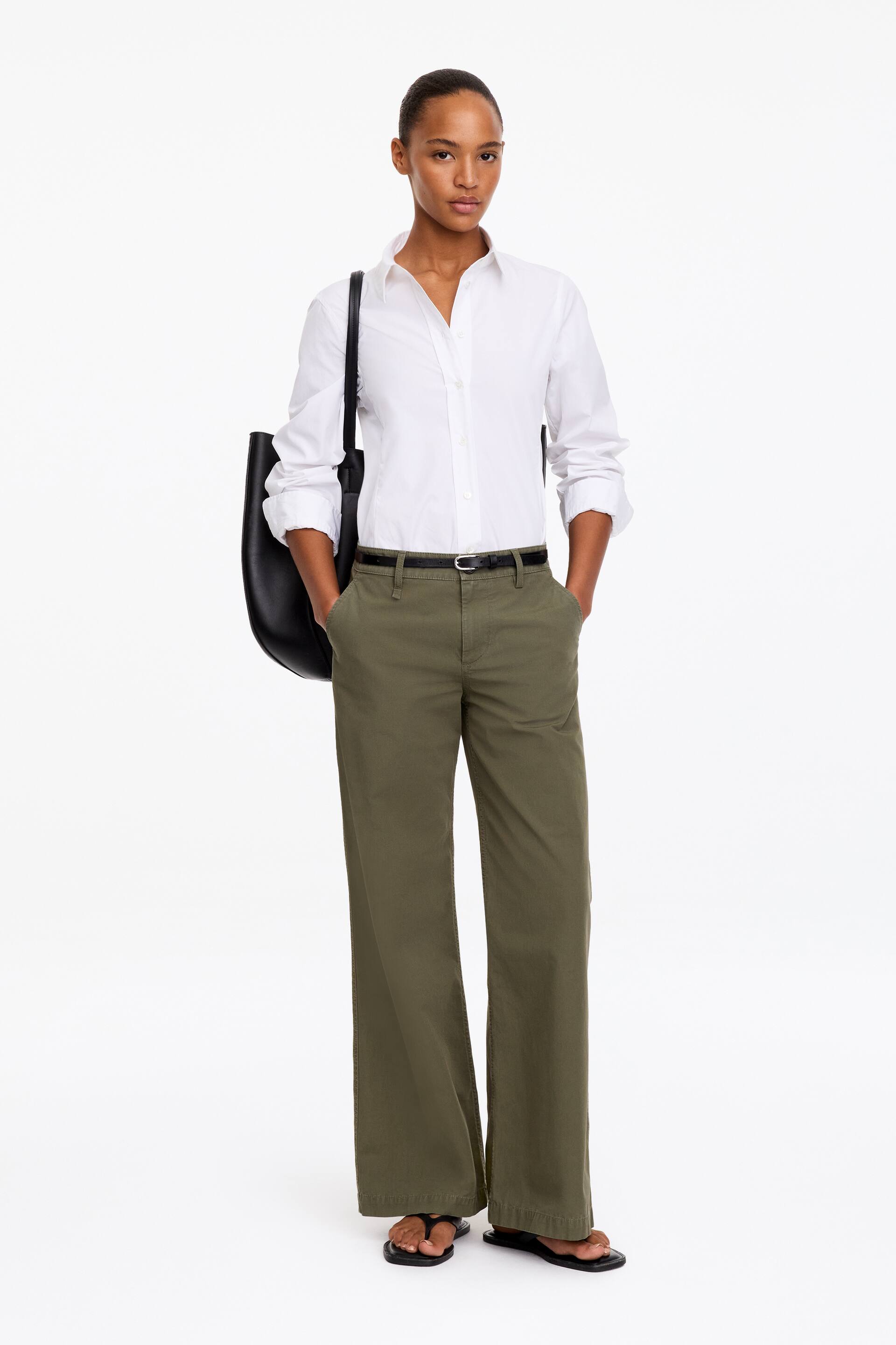 Visualizza l'immagine ingrandita: La modella indossa pantaloni chino a gamba larga verde oliva, con le mani inserite nelle tasche laterali oblique. I pantaloni a vita bassa sono chiusi da una sottile cintura nera e abbinati a una camicia bianca a maniche lunghe infilata dentro.